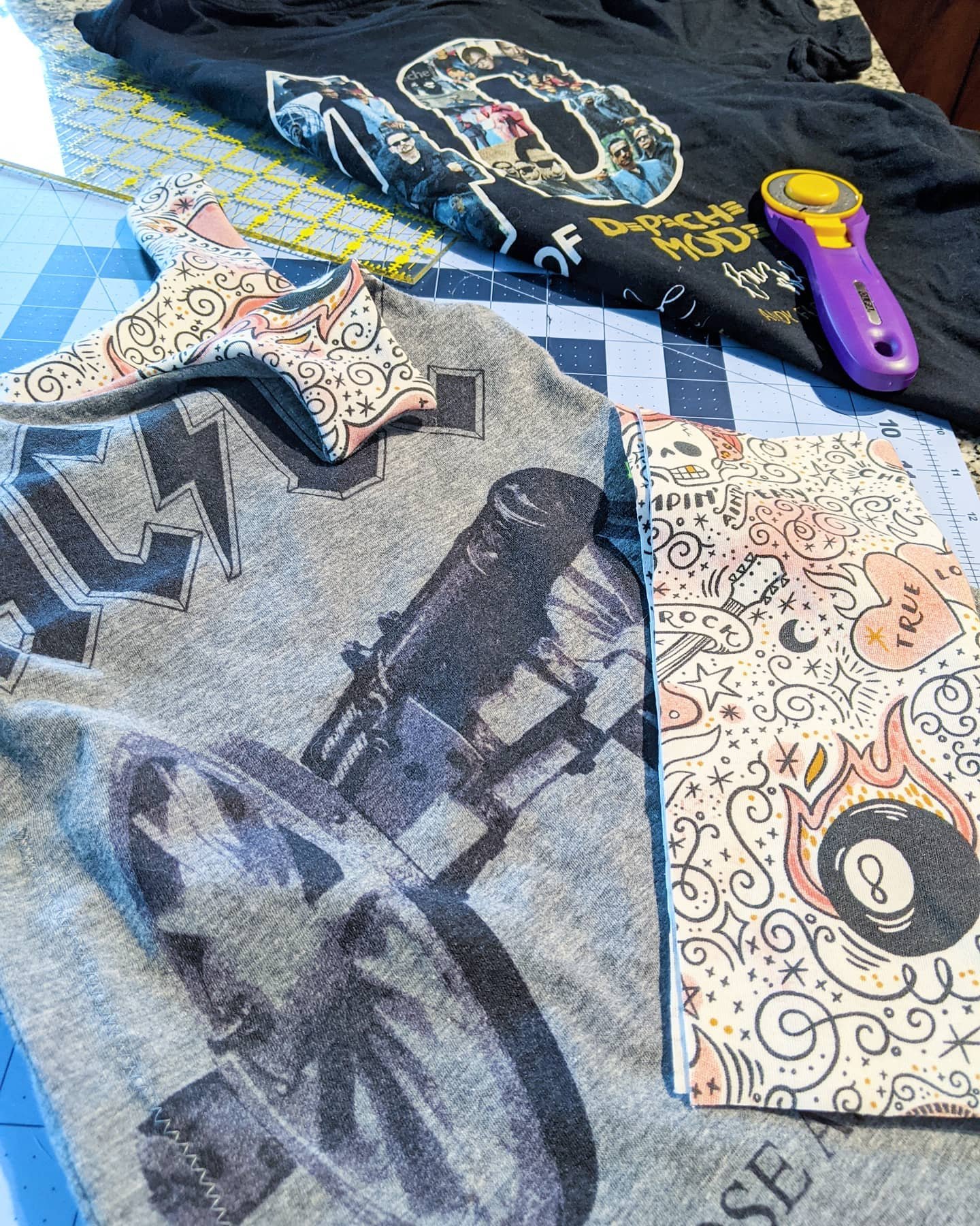 Fabric pieces, a T-shirt with a motorcycle print, colorful patterned fabric with skull and heart designs, a rotary cutter, and a cutting mat on a worktable.