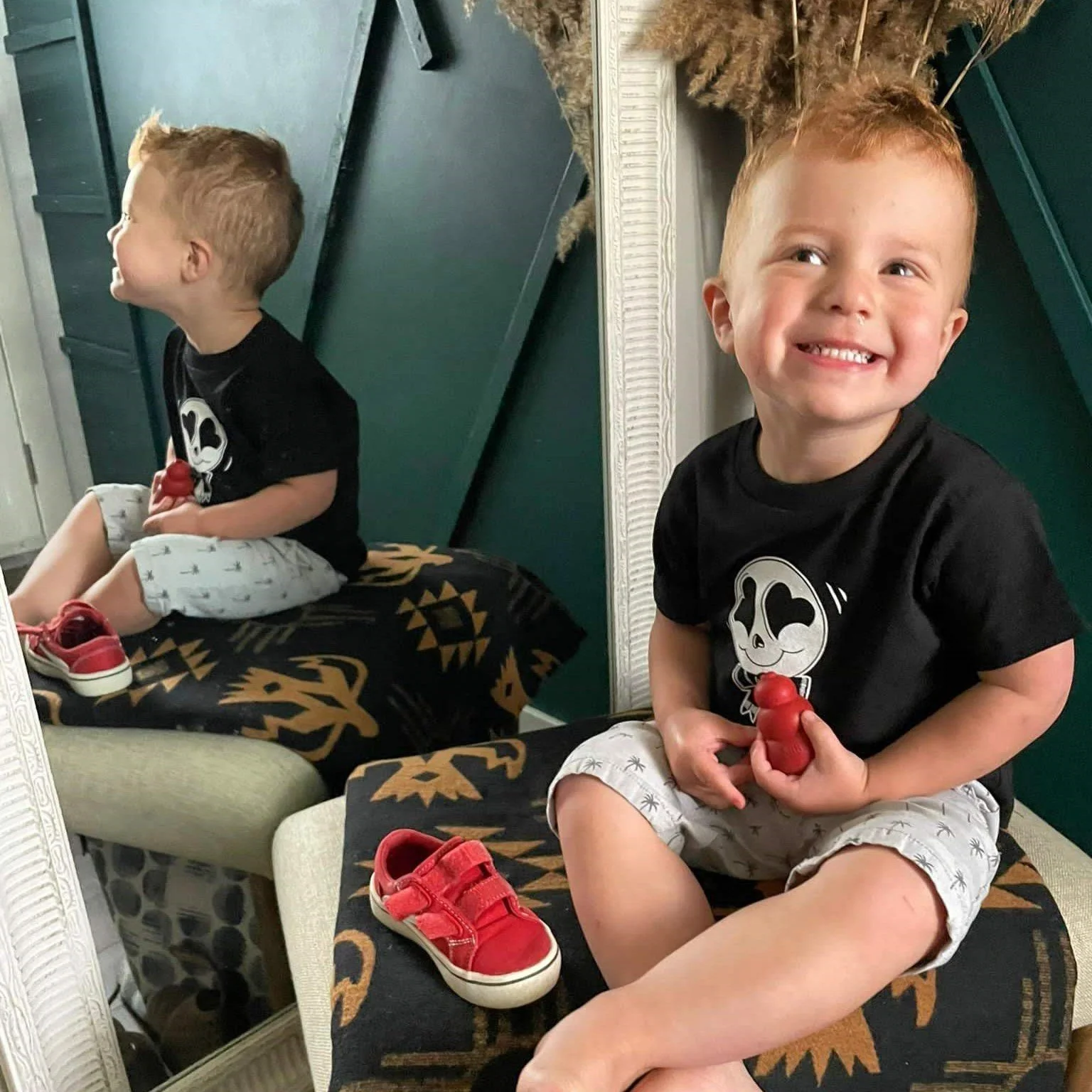 A young boy with red hair and a big smile sitting on a patterned ottoman, holding a red toy. He is wearing a black T-shirt with a cartoon character and light-colored shorts. There is a mirror behind him reflecting his side profile, and a pair of red sneakers is on the ottoman next to him.