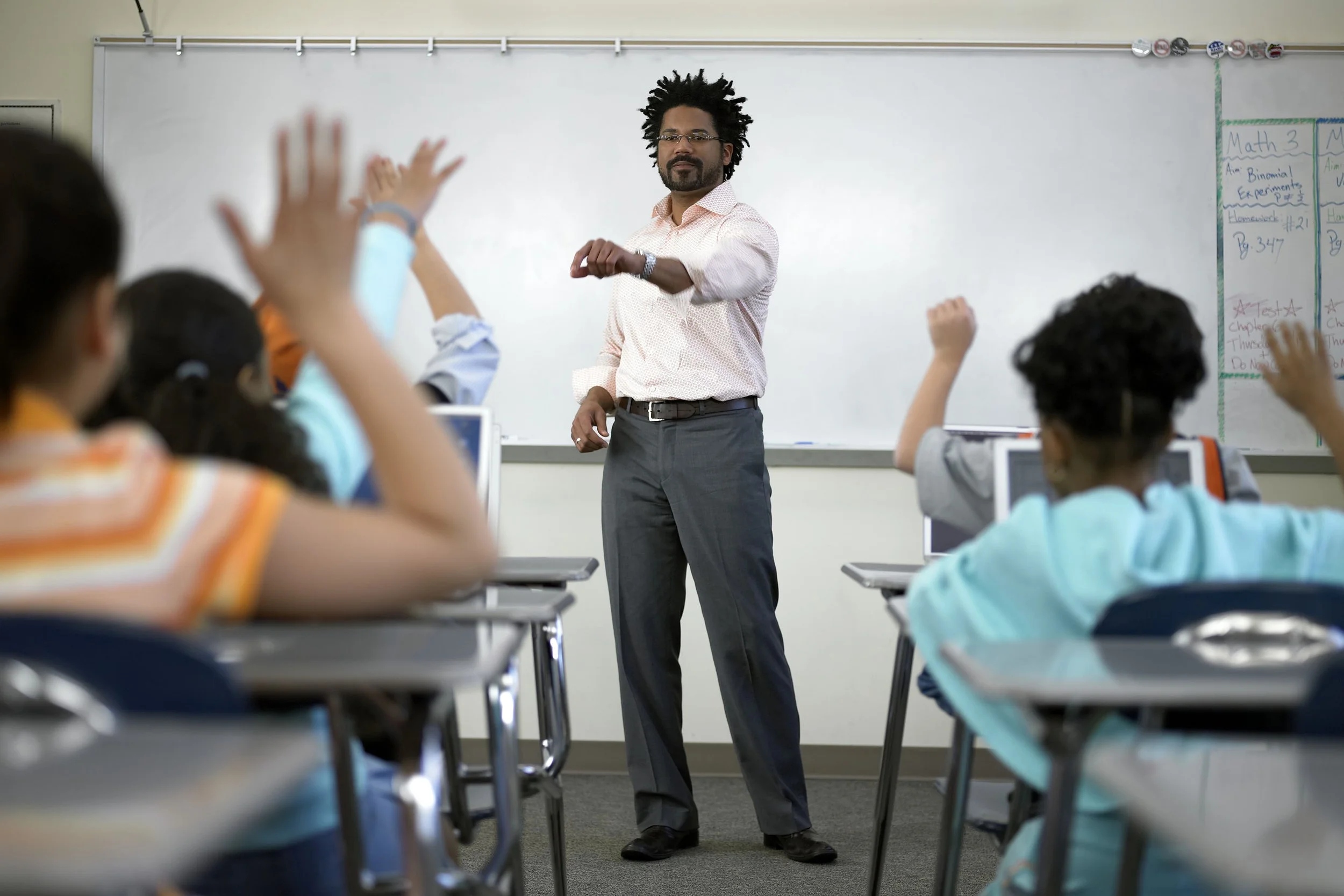 A male teacher in a classroom standing in front of a whiteboard, with students sitting at desks raising their hands.