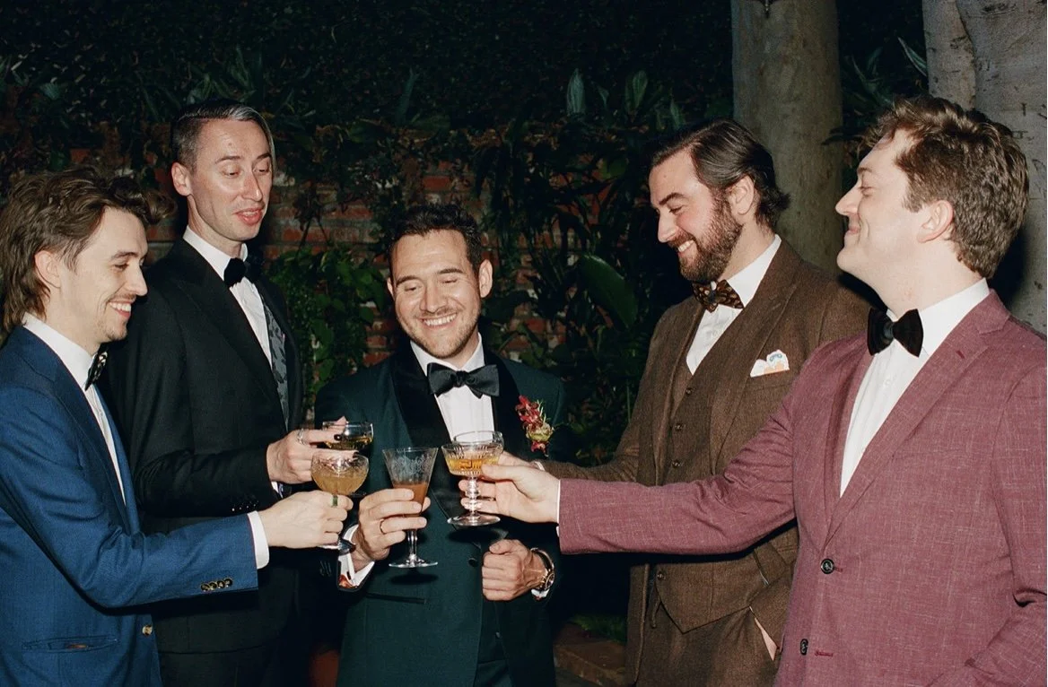 Group of six men dressed in suits and tuxedos smiling and holding cocktails at a celebration or party.