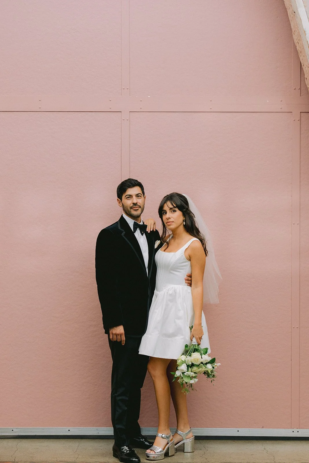 A newlywed couple stands arm in arm against a pink wall, with the woman holding a bouquet of white flowers and wearing a white dress with chunky heels, and the man in a black tuxedo and bow tie.