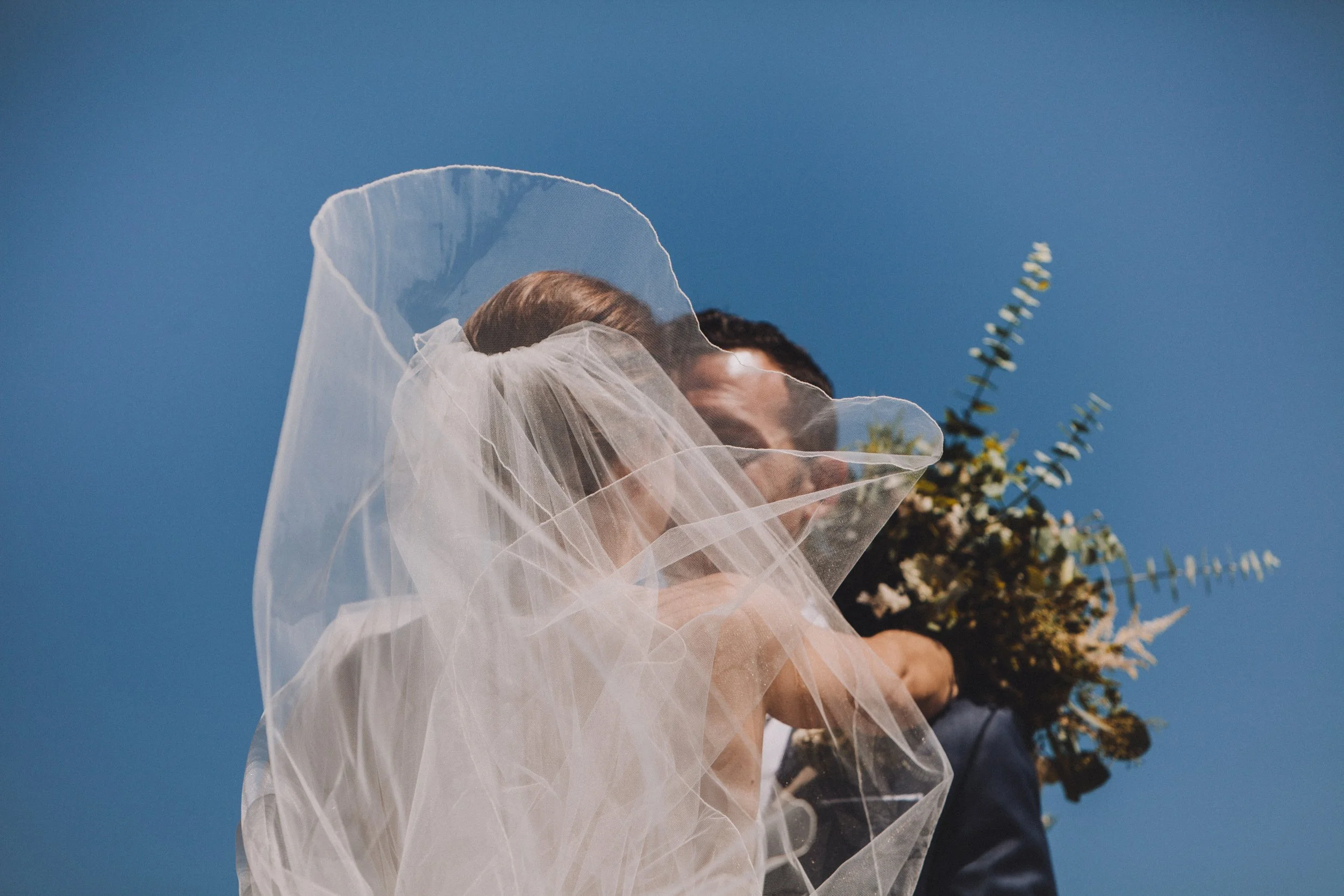 Wedding dress, a pair of white high-heeled shoes, a glass of water, and some decorative fabric on a white marble countertop with a brick wall background captured on 35mm film.