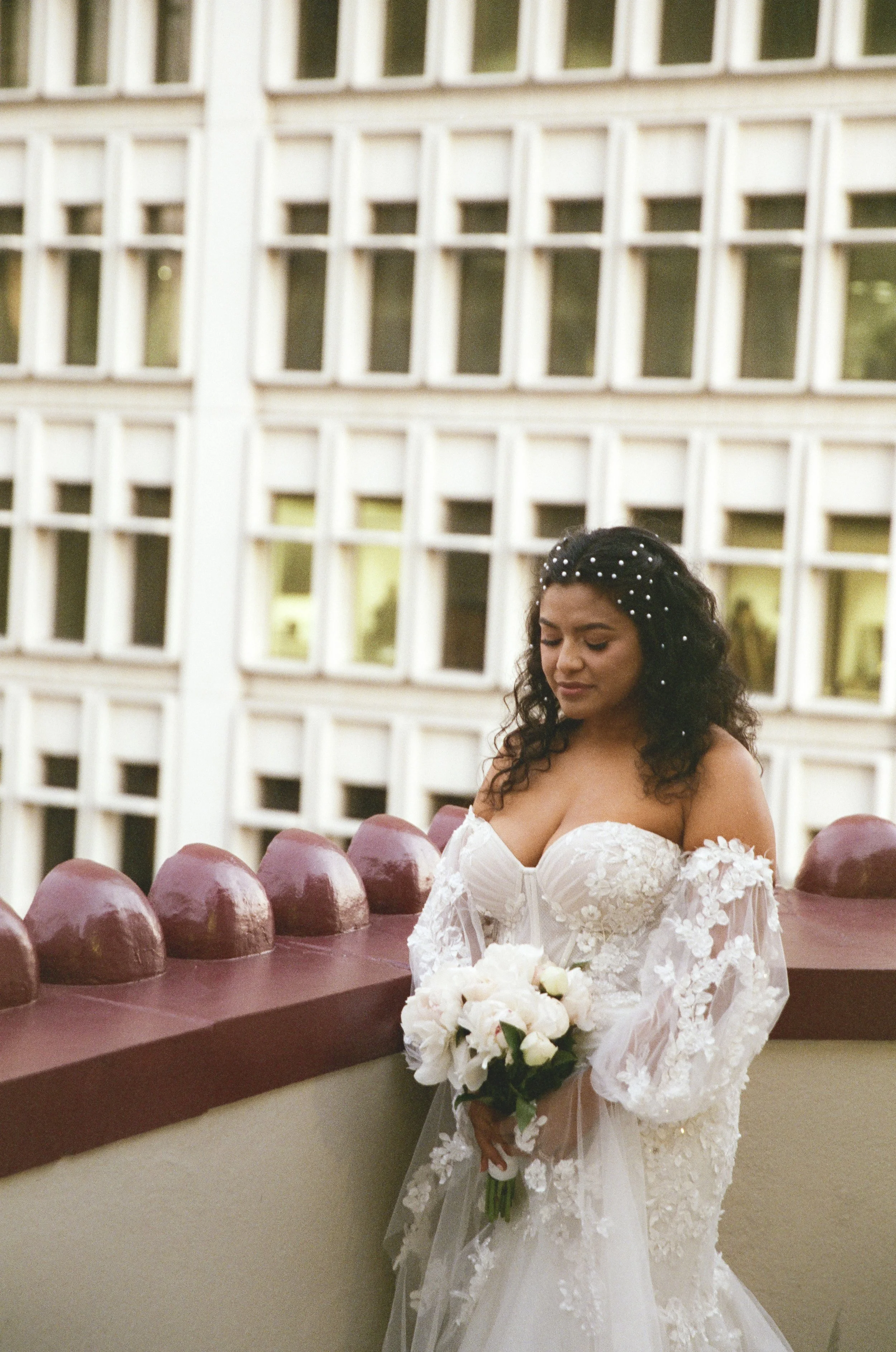 A bride in a white lace wedding dress holding a bouquet of white flowers outdoors, standing near a brown wall with rounded decorative features, in front of a building with multiple rectangular windows. She has dark, curly hair decorated with small wh