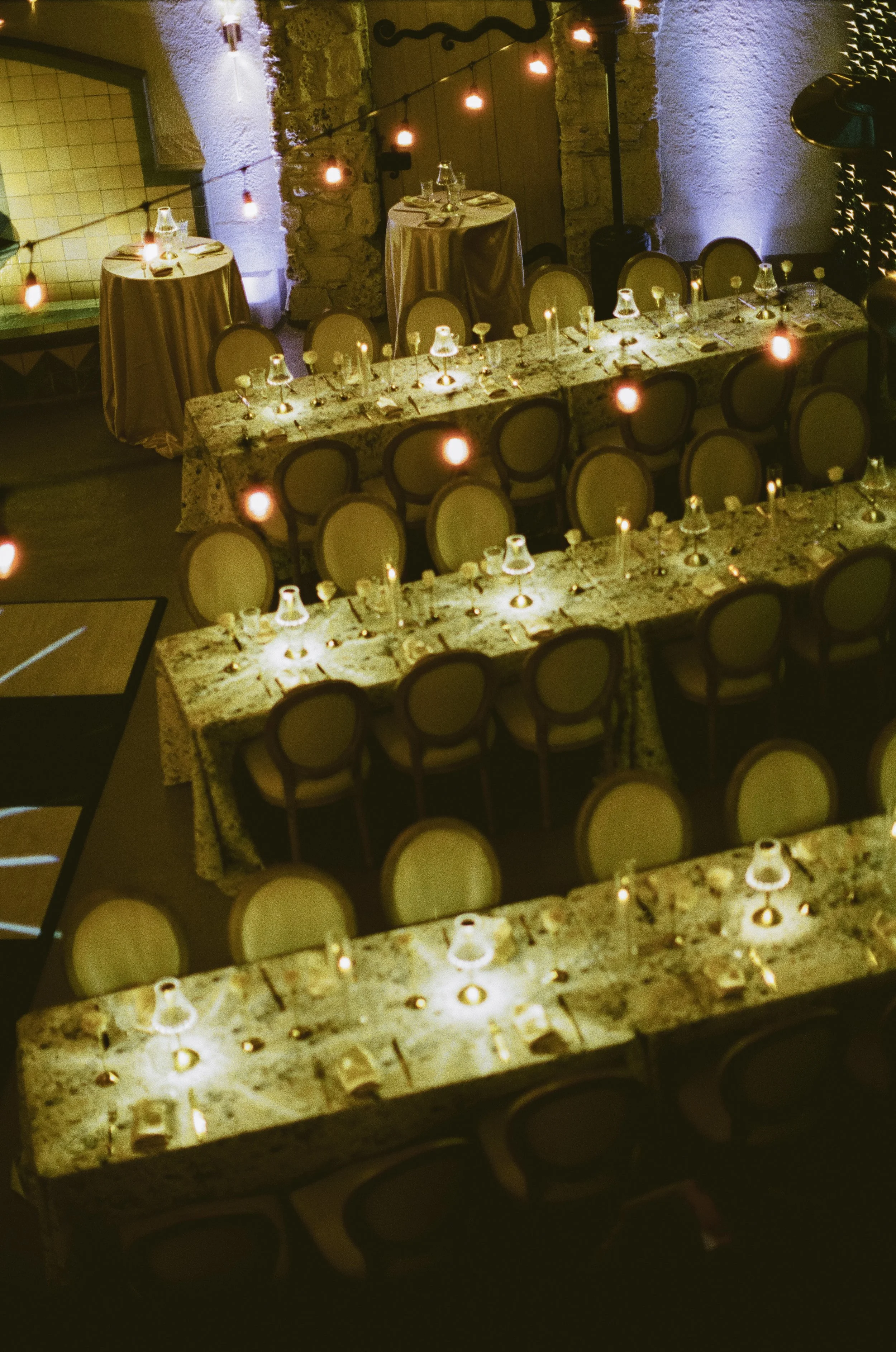 Evening dining setup with long marble tables, beige padded chairs, candlelit glasses, and string lights in a rustic indoor setting.