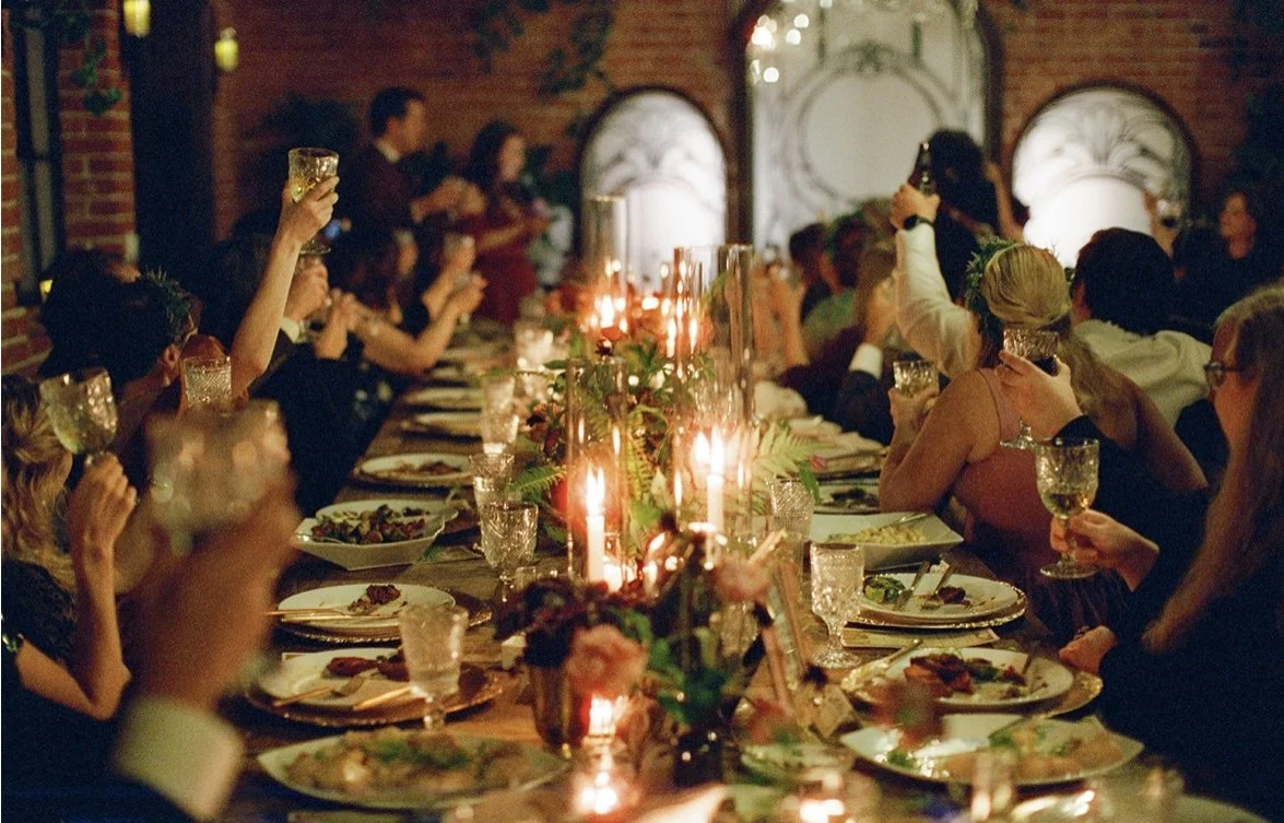 People seated at a long dining table raising glasses in a celebration, with candles, flowers, and elegant tableware in a warmly lit venue.