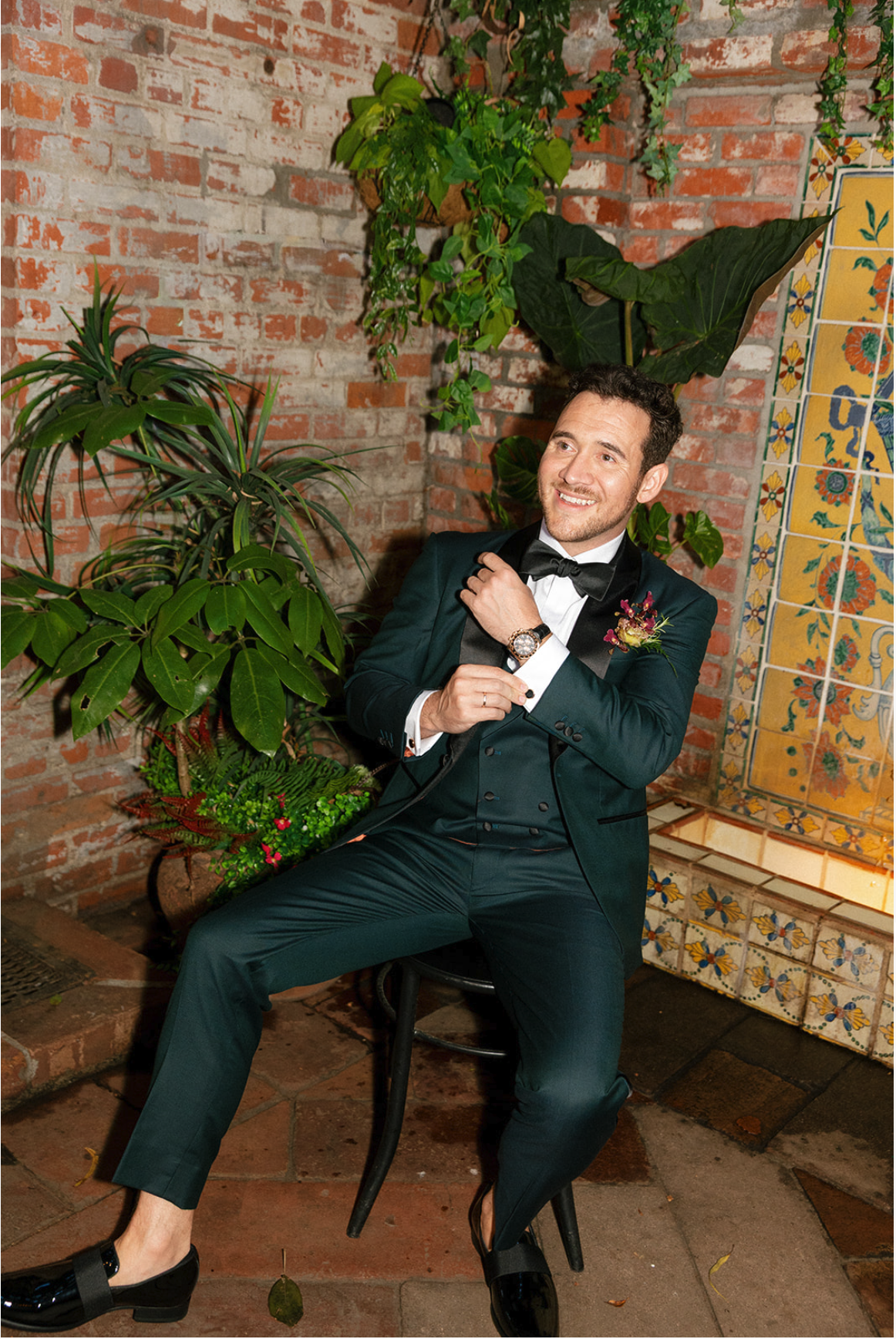 A smiling man in a tuxedo sitting on a black chair, adjusting his bow tie, with a brick wall and green plants in the background.