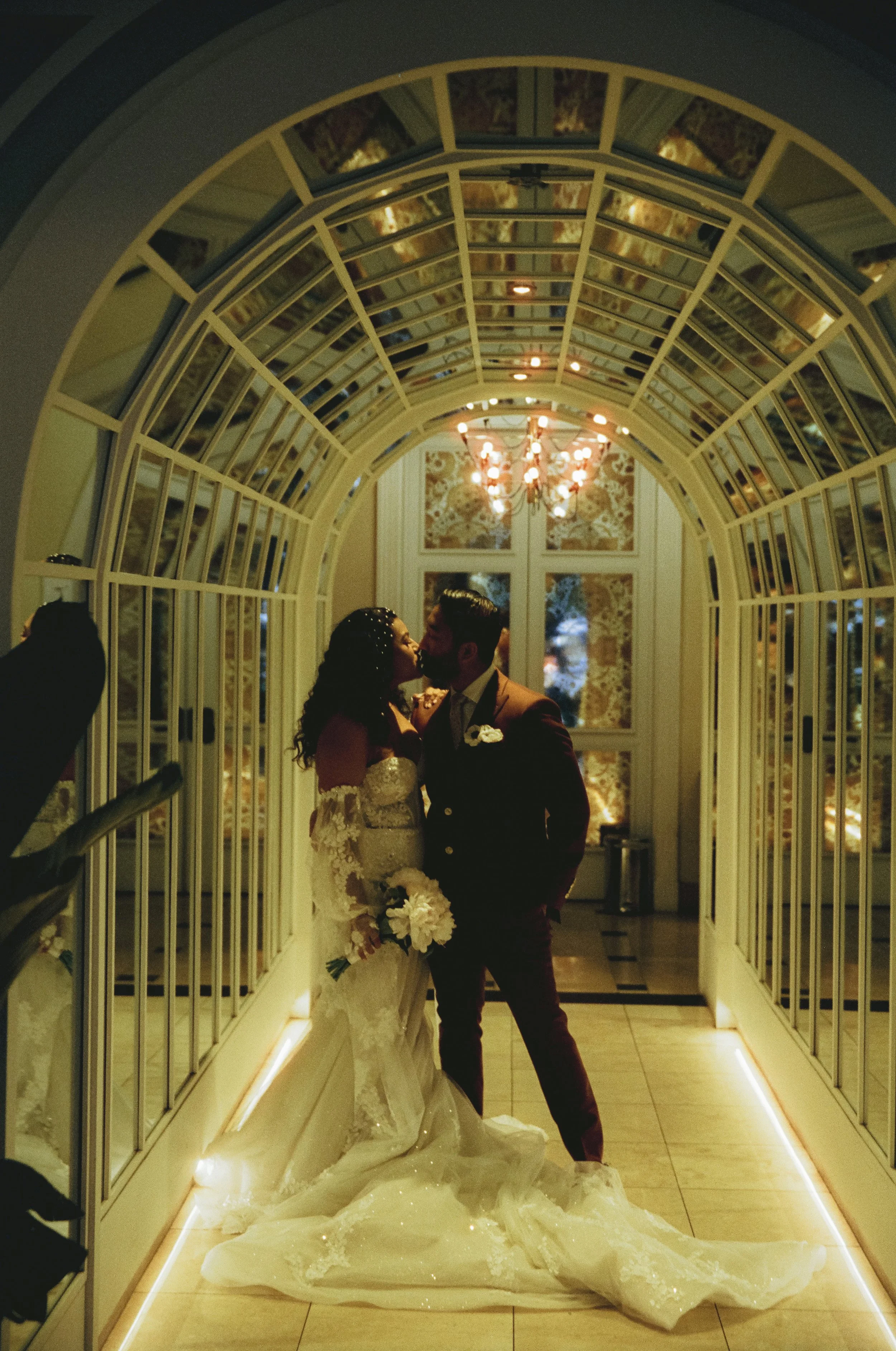 A newlywed couple sharing a kiss under a lit chandelier, silhouette in a decorative hallway with a glass archway and floral wallpaper at night.