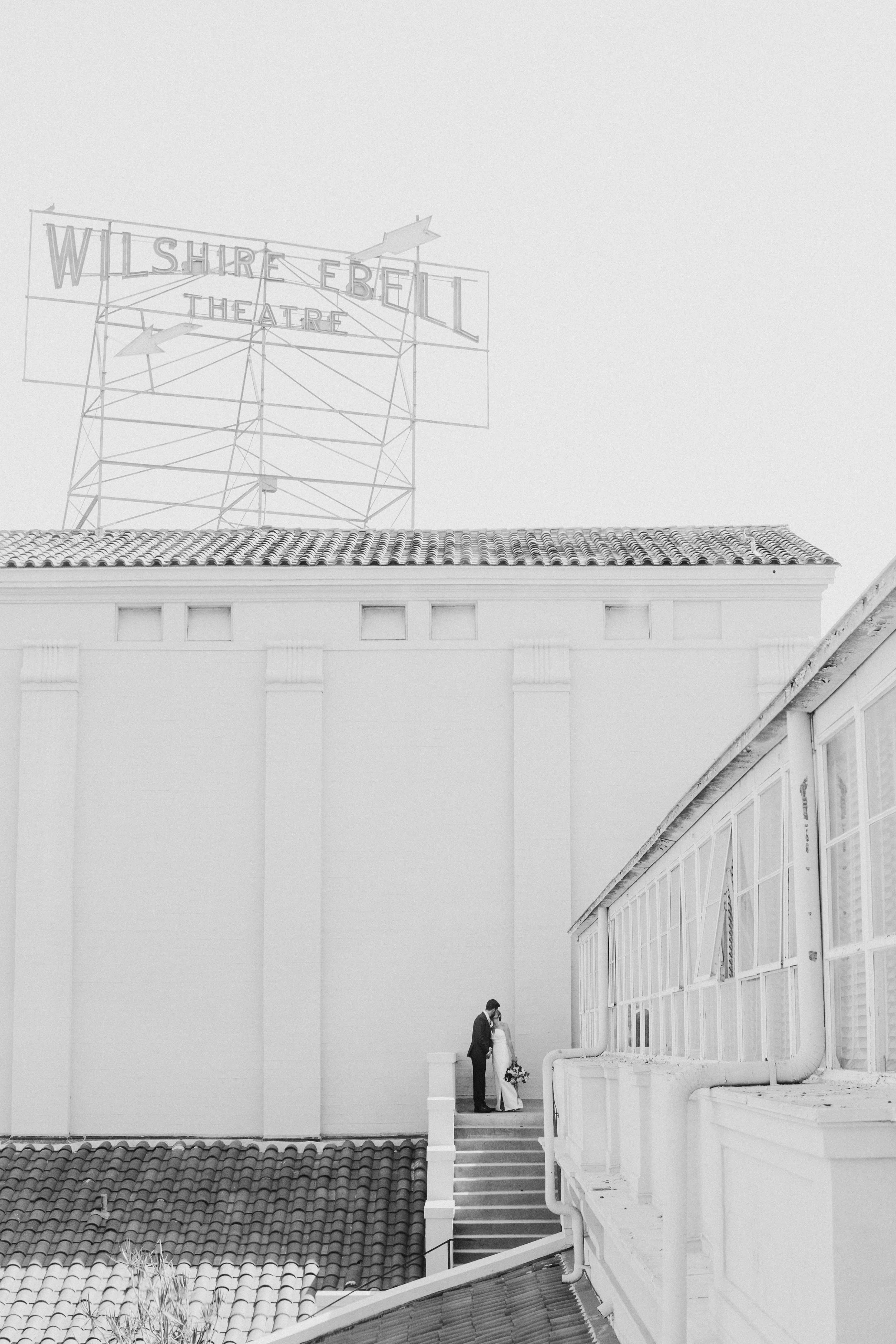 Black and white photo of the Ebell Los Angeles courtyard with a statue on a pedestal, surrounded by chairs and trees, with buildings and towers in the background.