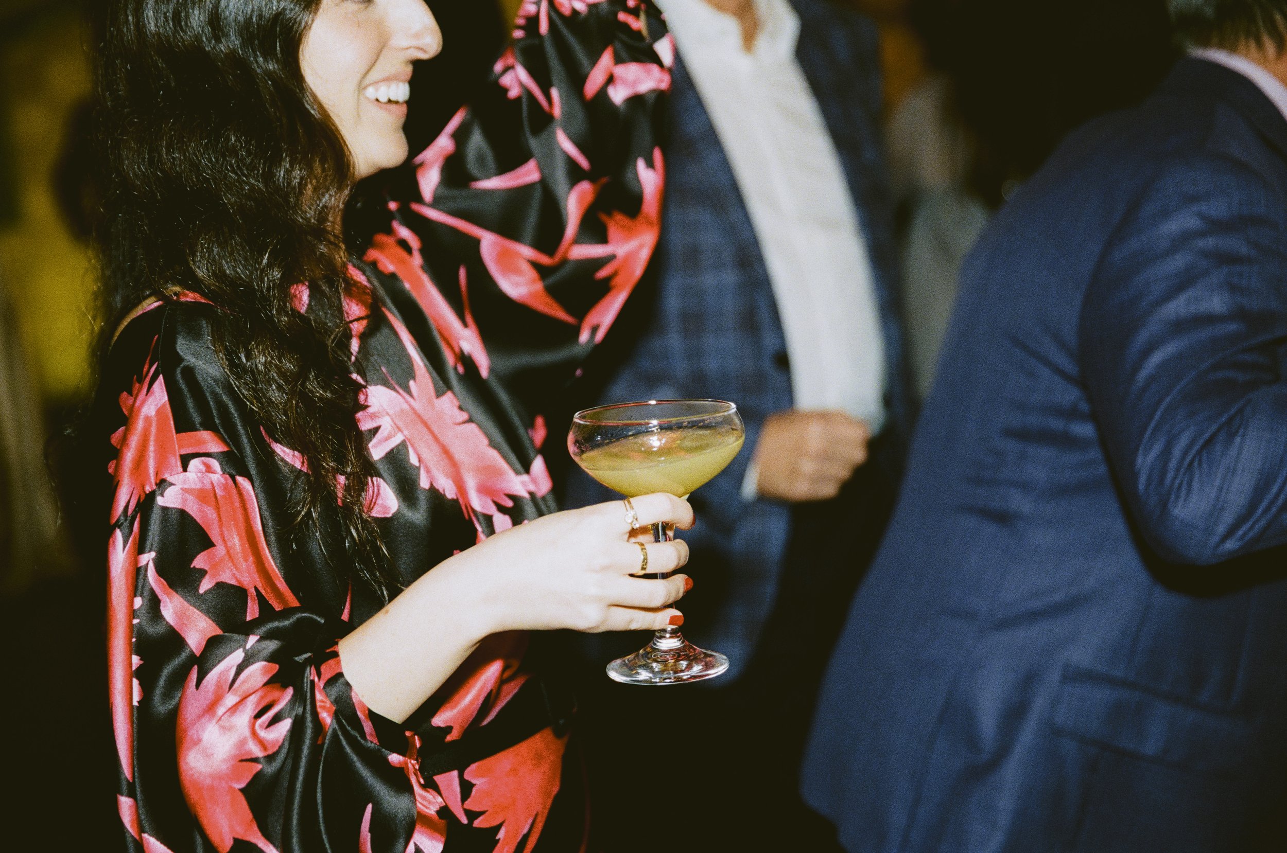 A woman holding a cocktail glass at a social event, dressed in a black and pink floral satin dress, smiling.