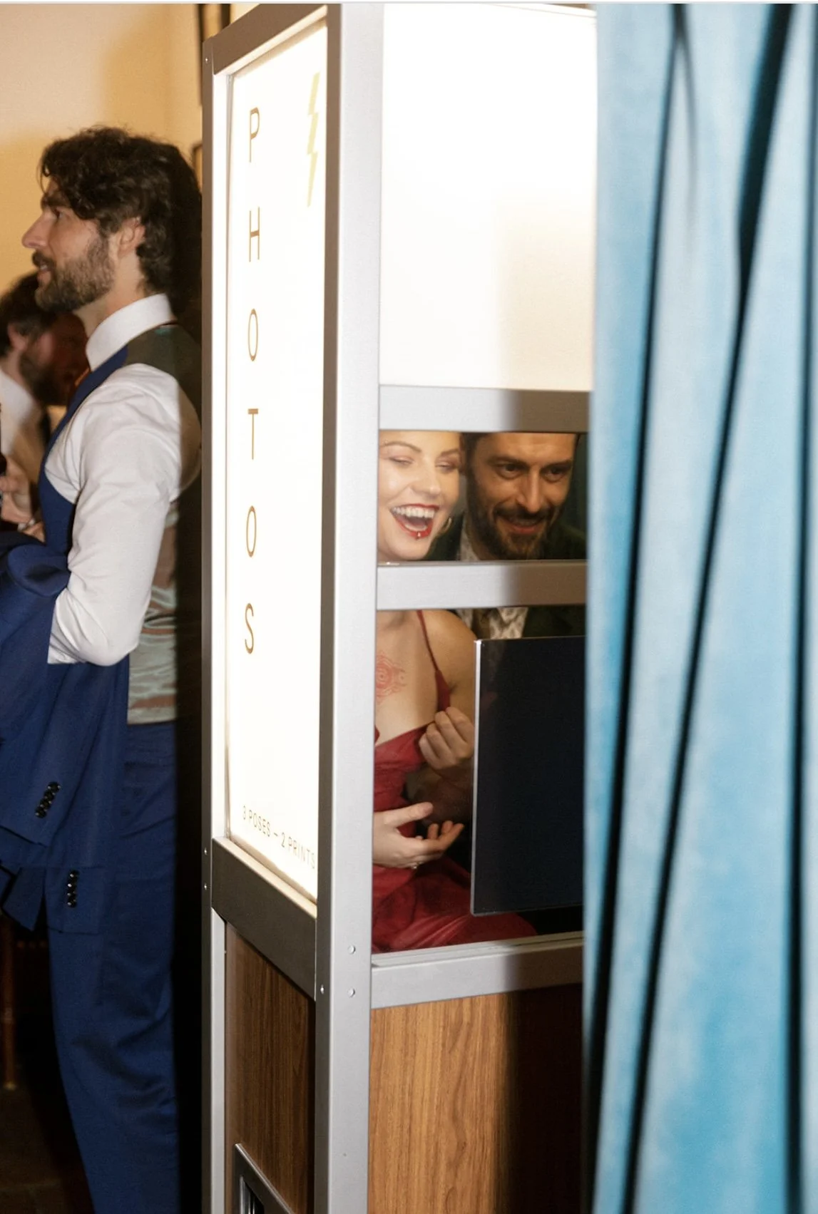 Two people, a woman in a red dress and a man, are smiling and looking at a screen inside a photo booth. They are partially visible through the booth's privacy divider.