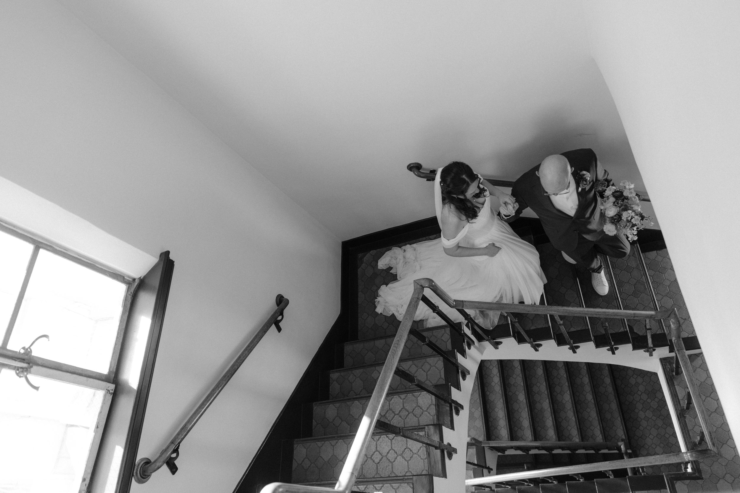 Black and white photo of a woman in a wedding dress and veil looking into a mirror while a person adjusts her hair or makeup.