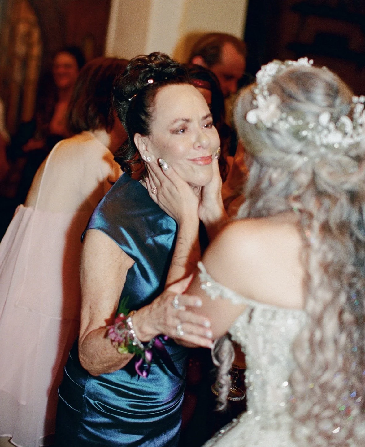 An emotionally moved woman in a blue satin dress holding face of another woman in a white wedding gown, sharing a tender moment at a wedding reception.