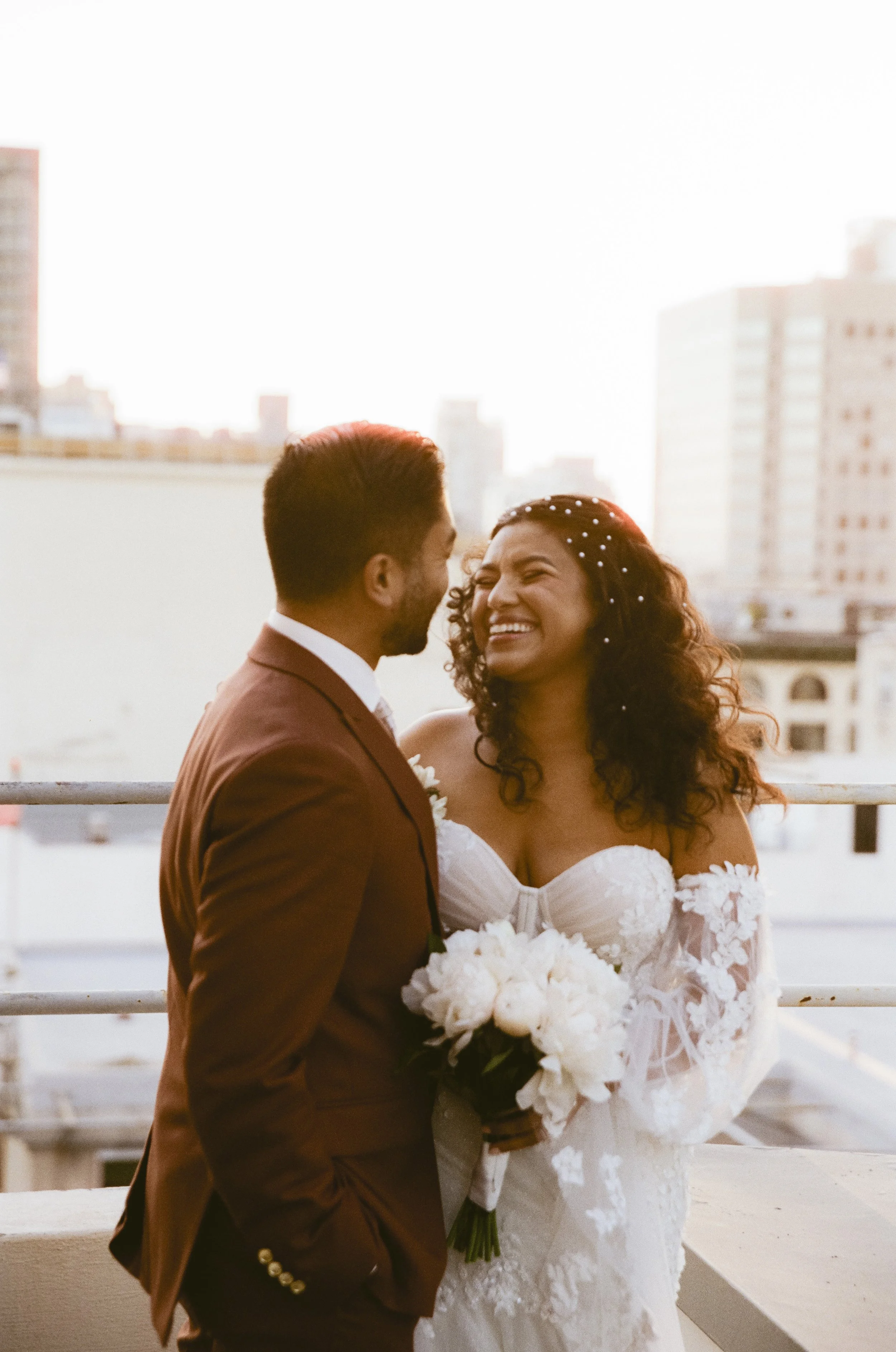 A newlywed couple stands on a rooftop, smiling and laughing, with a city skyline in the background during sunset. The bride wears a white gown with lace details and holds a bouquet of white flowers. The groom is dressed in a brown suit.