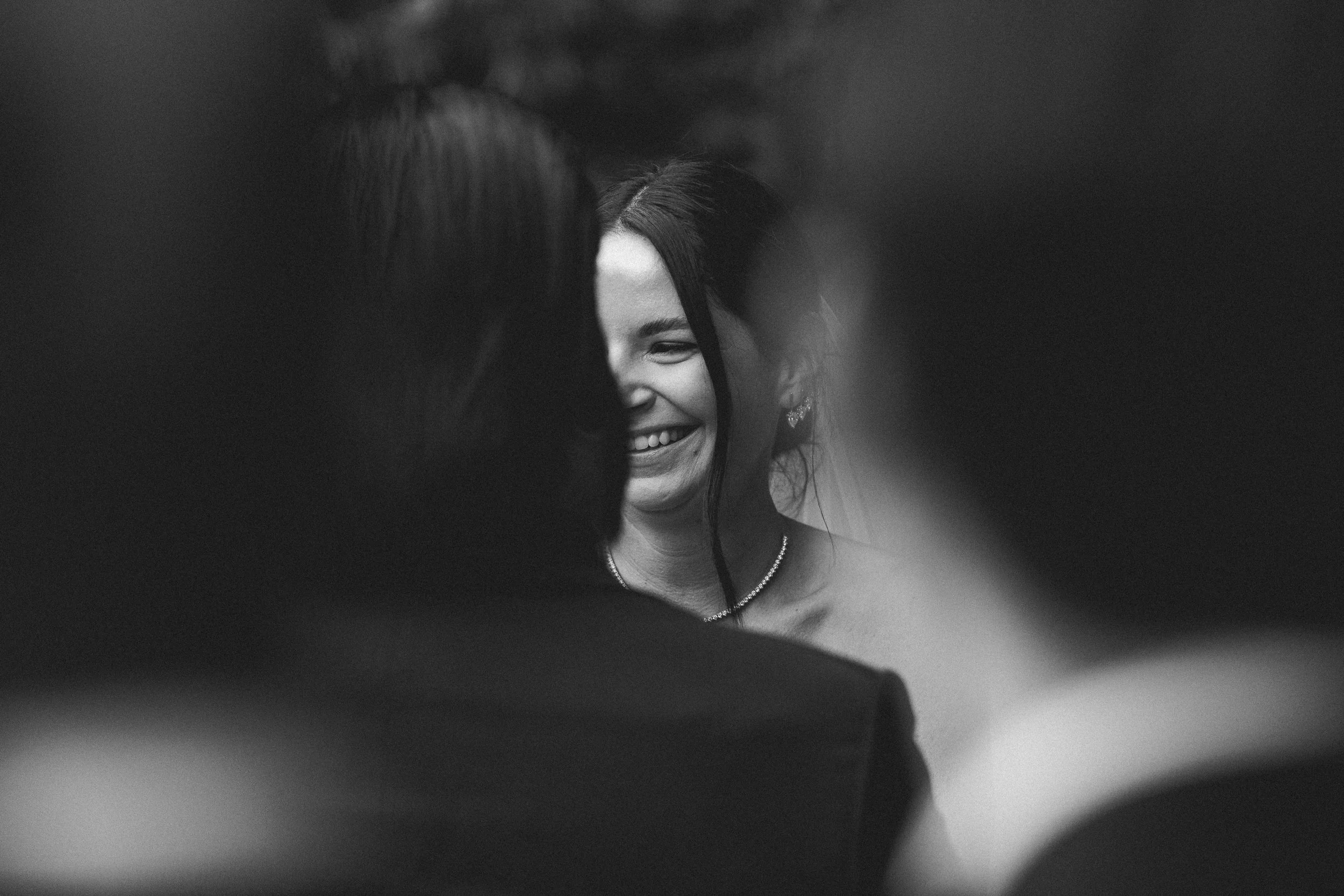 Black and white photo of a smiling woman with dark hair, wearing earrings and a necklace, surrounded by blurred figures at what appears to be a social event or gathering.