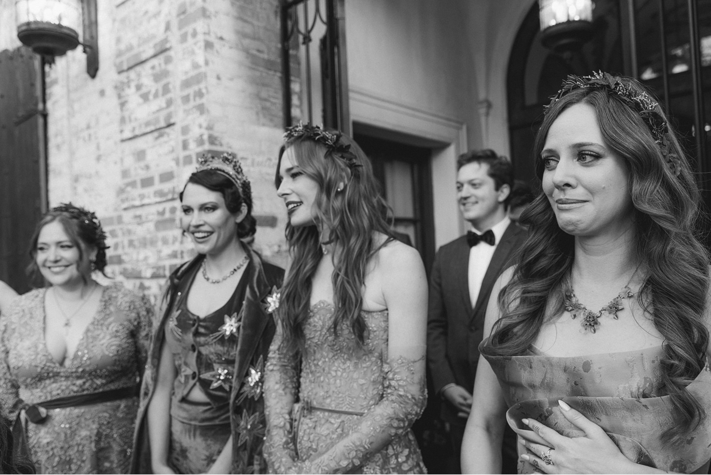 Group of women and a man dressed in formal attire at a social event, standing indoors near a brick wall.