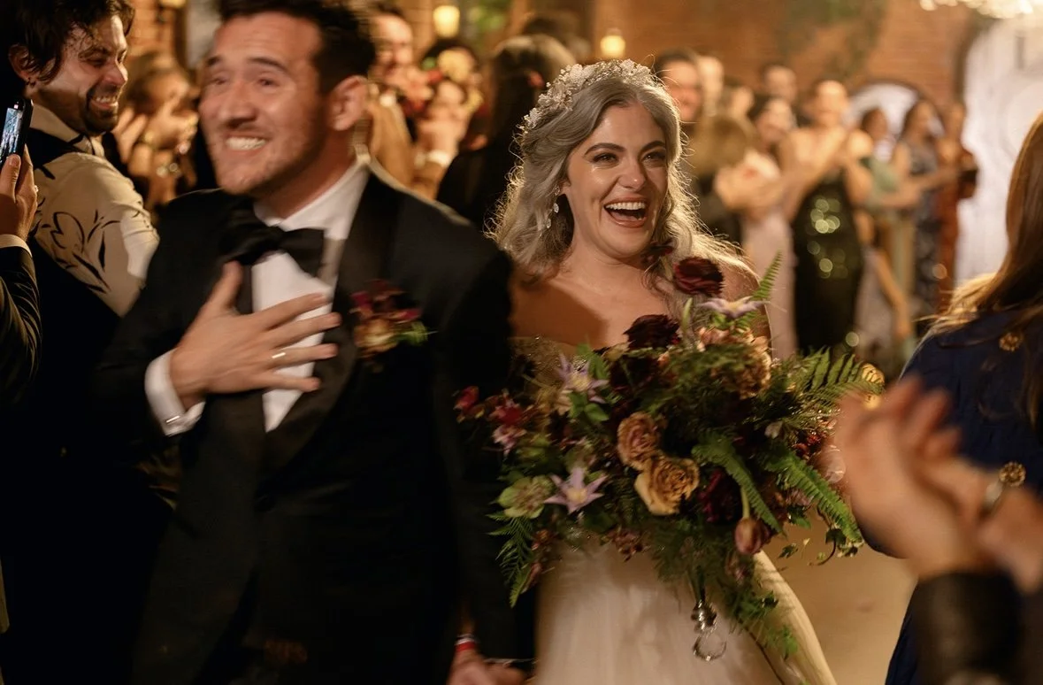 A happy bride in a wedding dress holding a large bouquet of flowers, smiling, and standing next to a man in a tuxedo, at a wedding celebration with guests in the background.