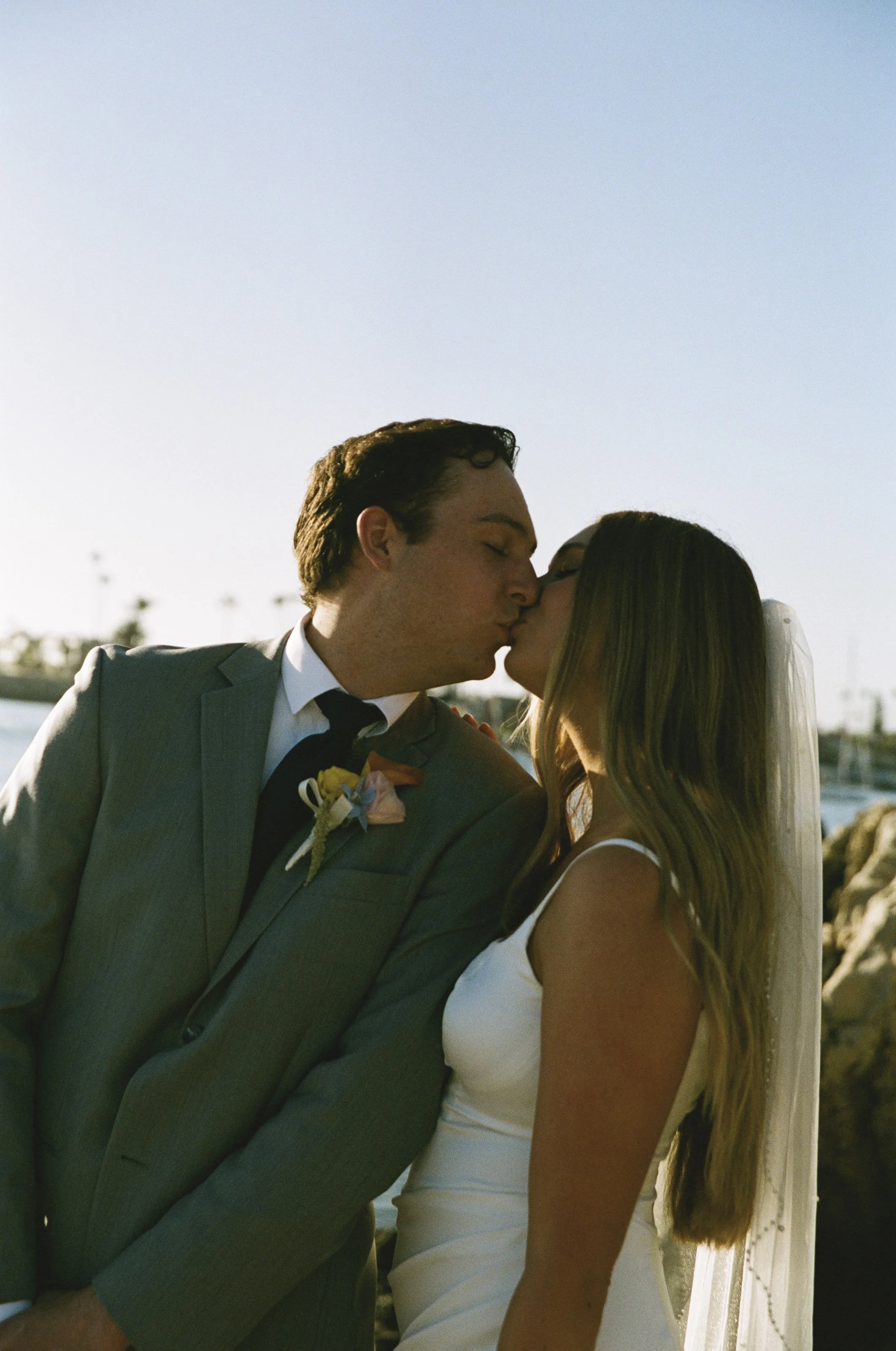 A couple in wedding attire sharing a kiss outdoors near the water during sunset.