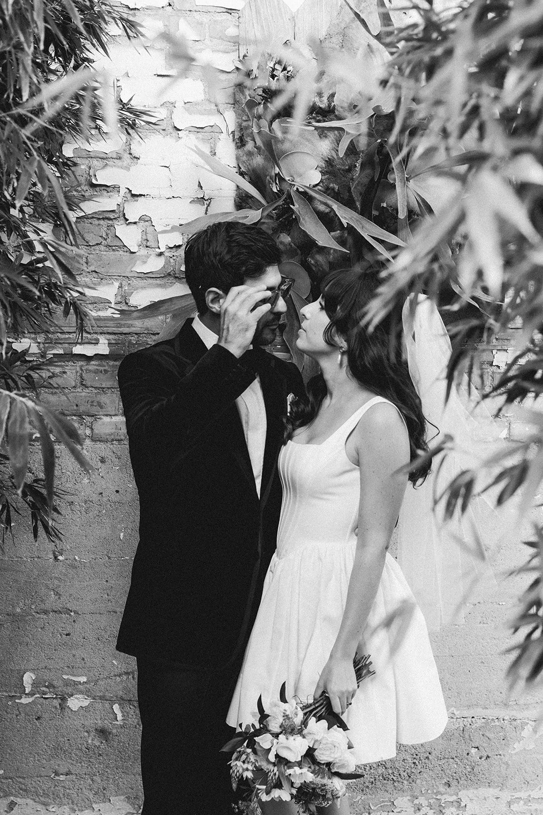A black and white photo of a couple standing close together against a brick wall surrounded by plants, gazing into each other's eyes, with the man adjusting his glasses and the woman holding a bouquet of flowers.