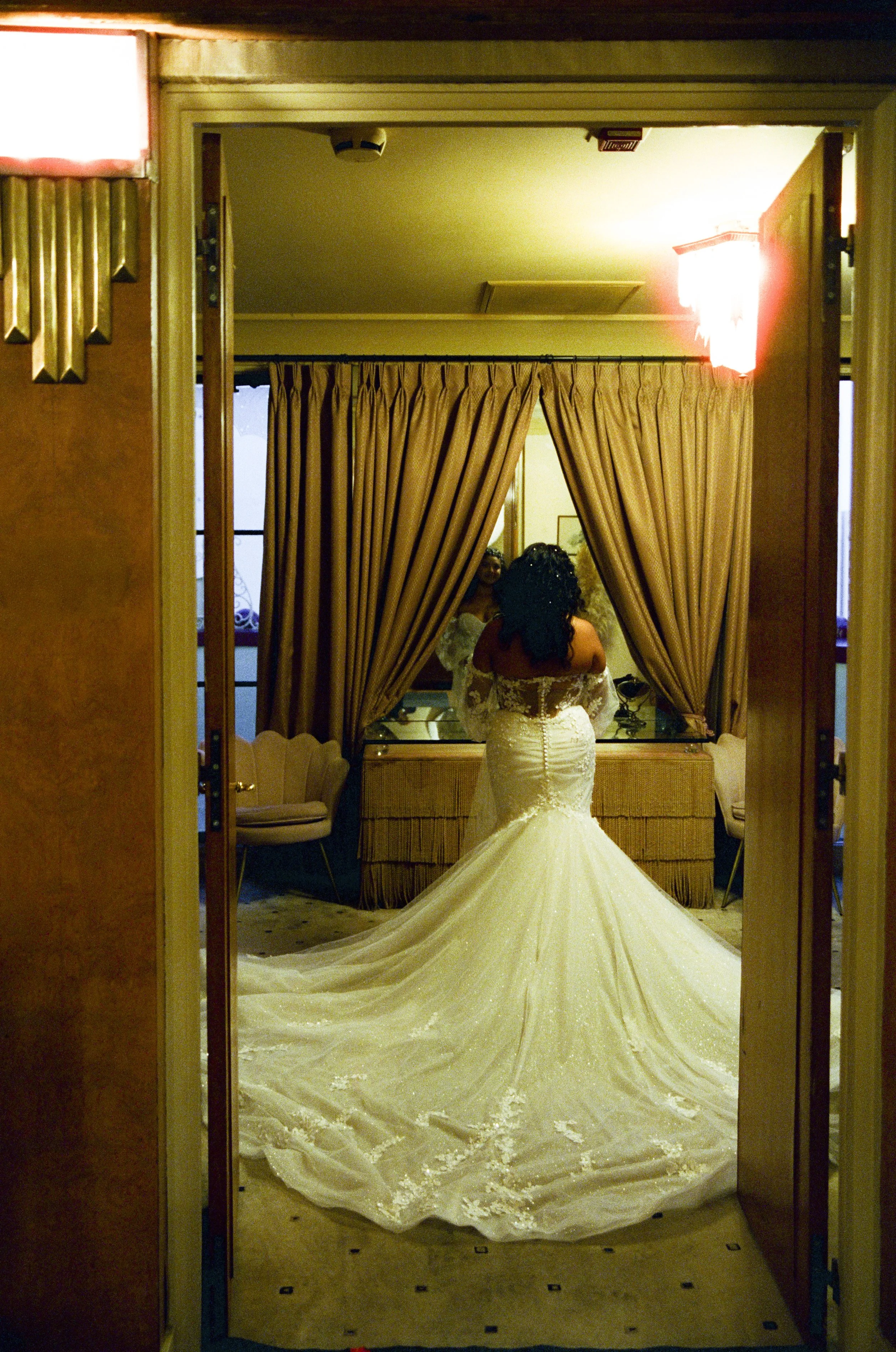 A bride in a white wedding dress with a long train stands in front of a mirror, seen from behind, as she gets ready in a room with gold curtains and vintage furniture.
