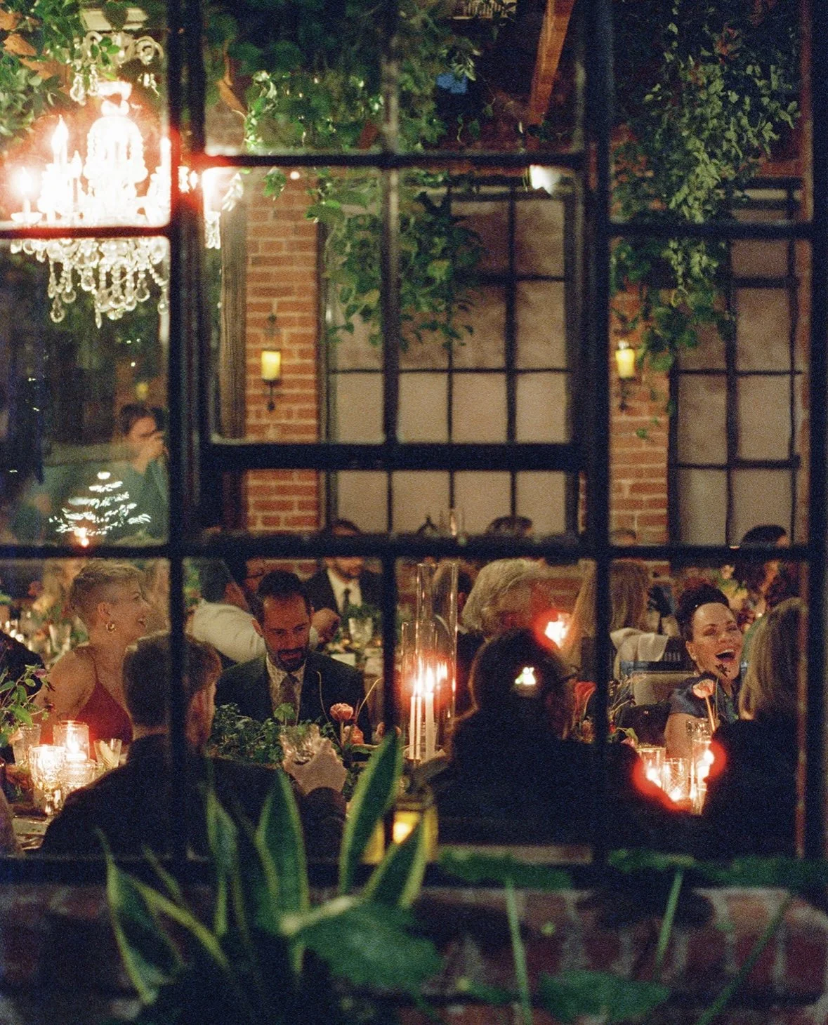 People gathered around a dining table at a festive event, seen through a window with black bars, candlelit setting, and greenery, with a chandelier and brick wall in the background.