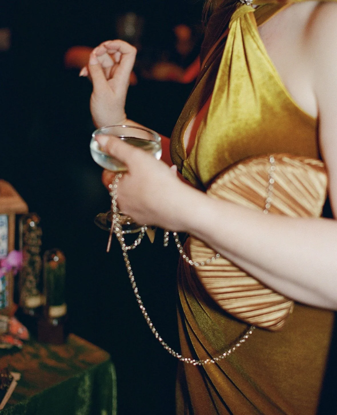 A woman wearing a shiny, two-tone gold and bronze dress holding a glass of champagne, with a pearl necklace hanging from her hand, and a blurred background with bottles and objects.