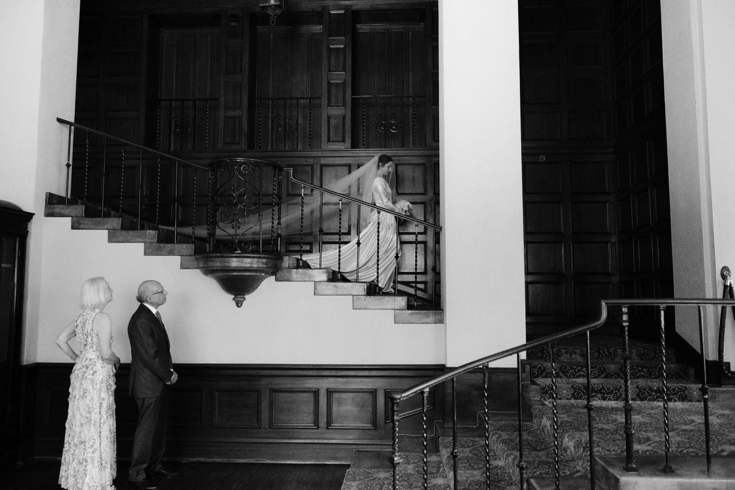 Black and white photo of a bride and groom holding hands outdoors, with the bride wearing heart-shaped sunglasses and a veil, and the groom in a tuxedo.