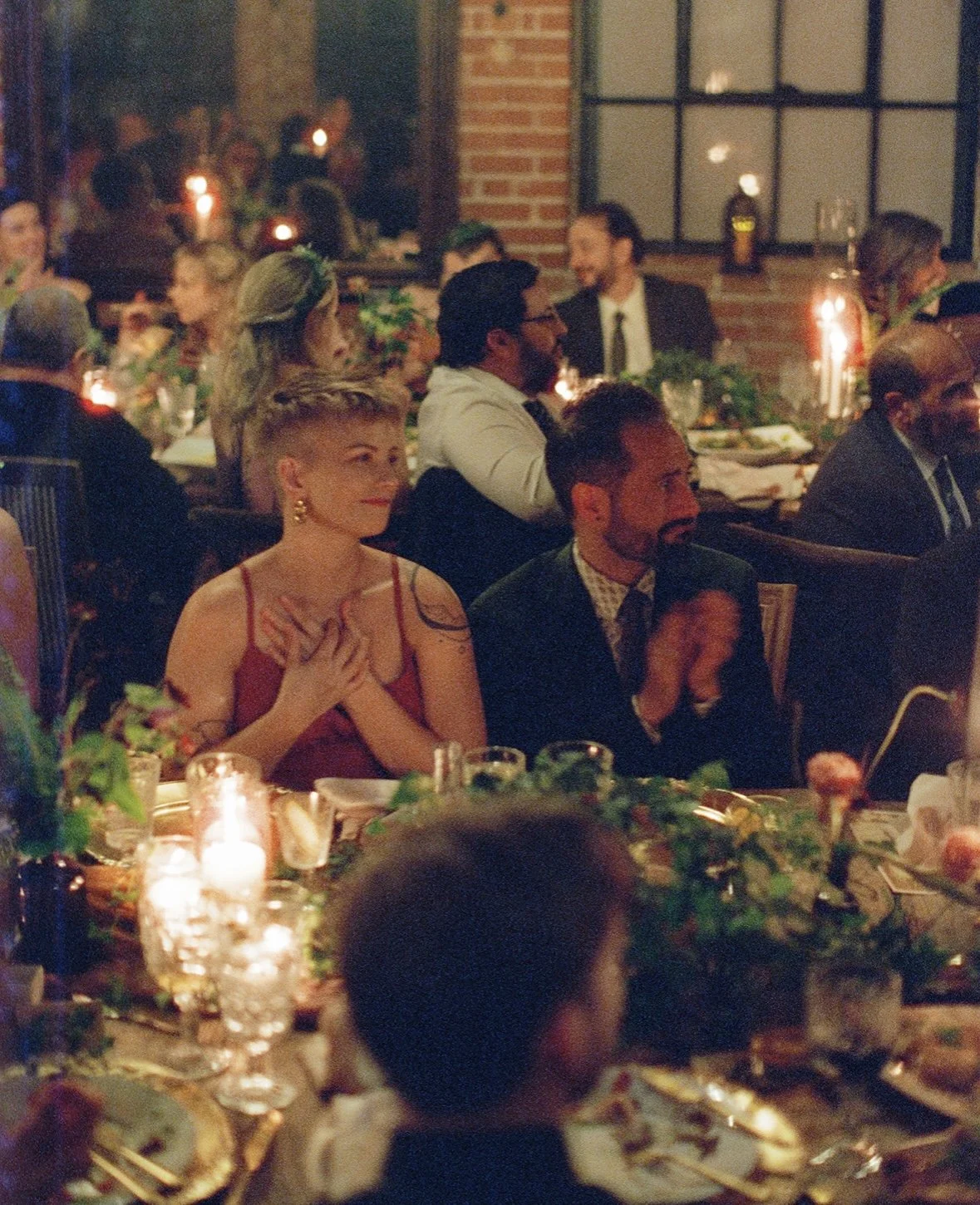 People attending a dinner event in a warmly lit, rustic-style restaurant with exposed brick walls and large windows, some clapping and enjoying the celebration.