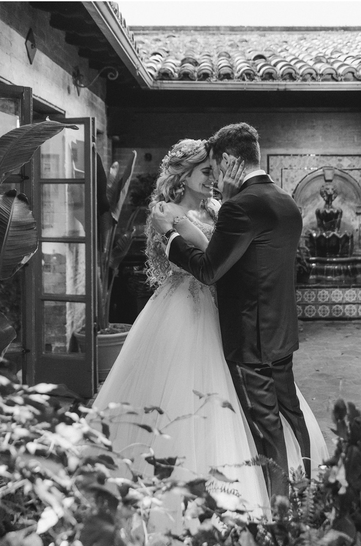 A black and white photo of a bride and groom dancing close together indoors, smiling with foreheads and hands touching, surrounded by plants and decorative art in the background.