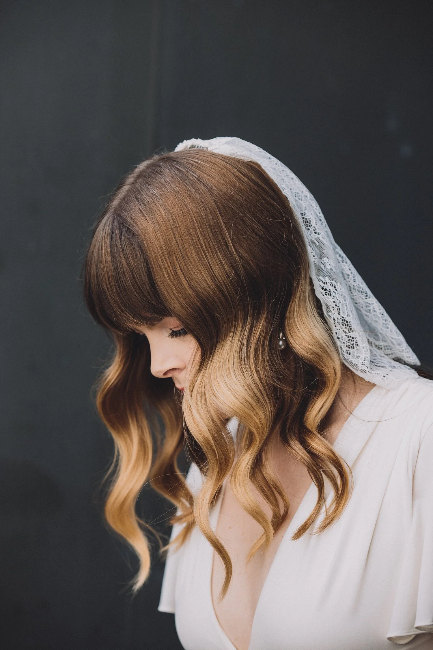A woman with long, wavy, brown hair and bangs wearing a white dress and a lace head covering, looking down against a black background.