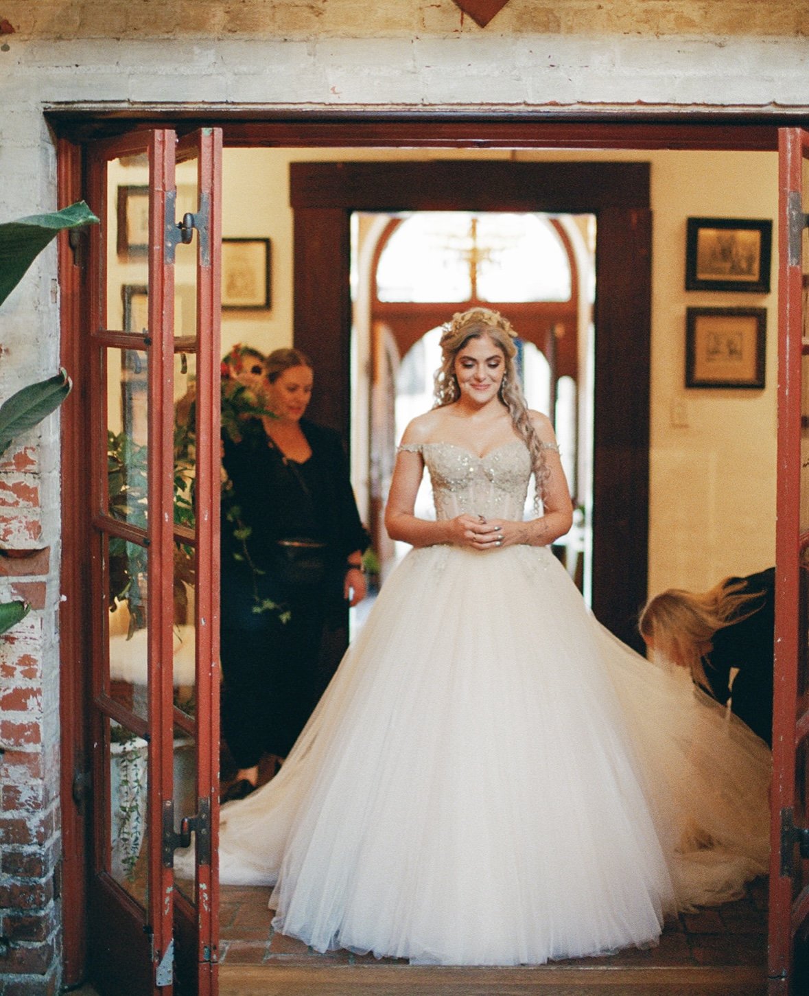 A bride in a white wedding gown smiling with her hands clasped, standing inside a house with two women helping with her dress, one adjusting the train and the other standing nearby. The house has warm lighting, wood accents, and framed pictures on the wall.
