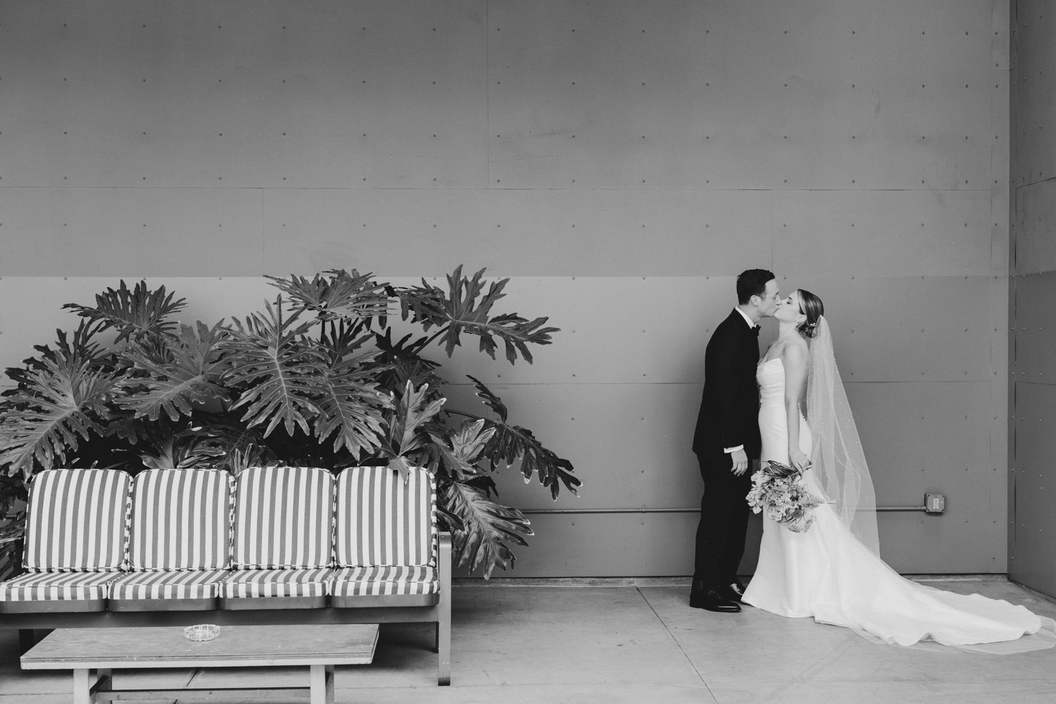 bride wearing strapess fitted dress with long train and long white veil kissing groom wearing classic black tux; couple standing in corner of clean patio of the Valentine Los Angeles, with just a striped patio chair on opposite corner