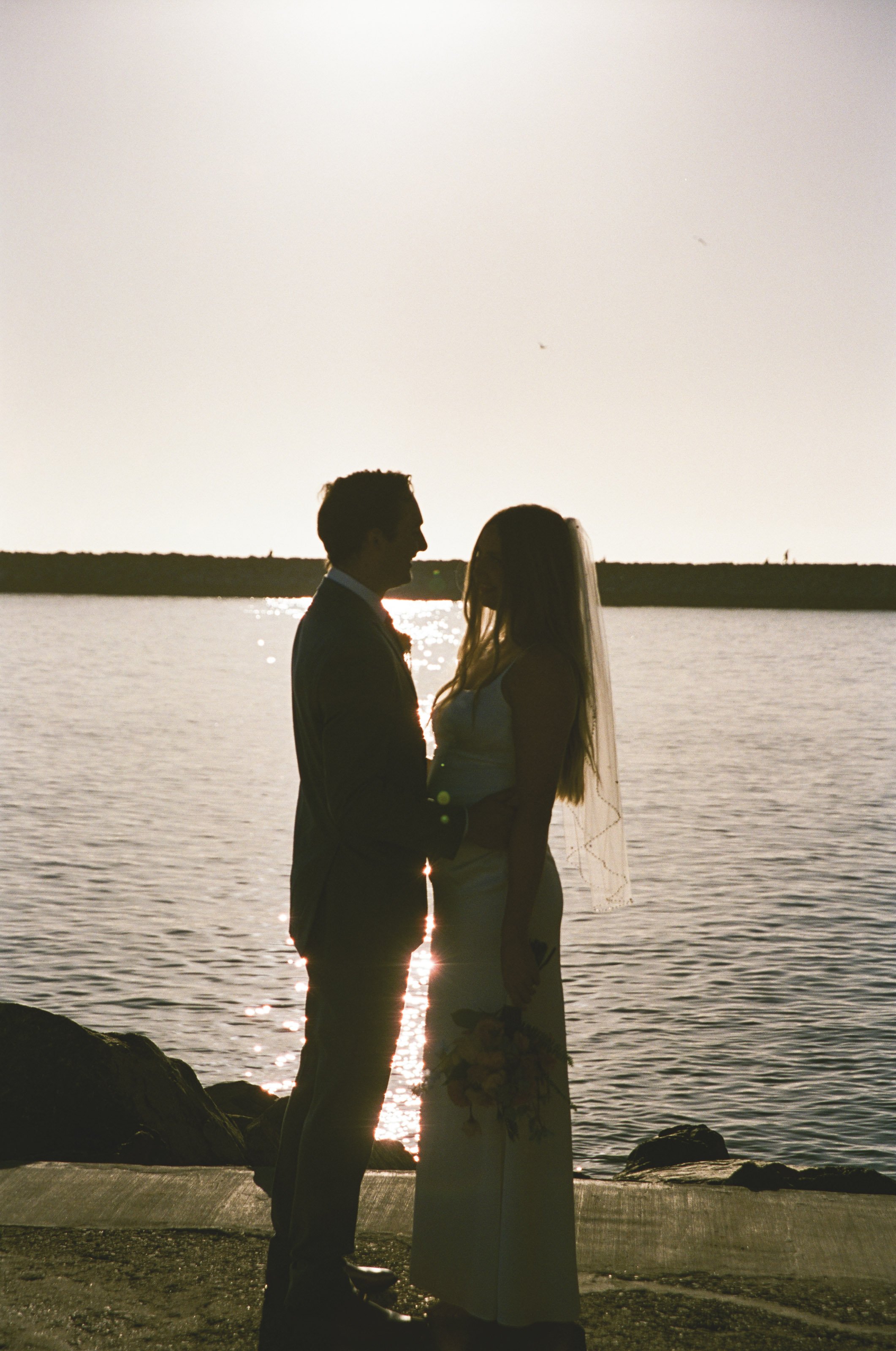 Silhouette of a bride and groom standing by a body of water during sunset, facing each other, with the sun reflecting on the water.