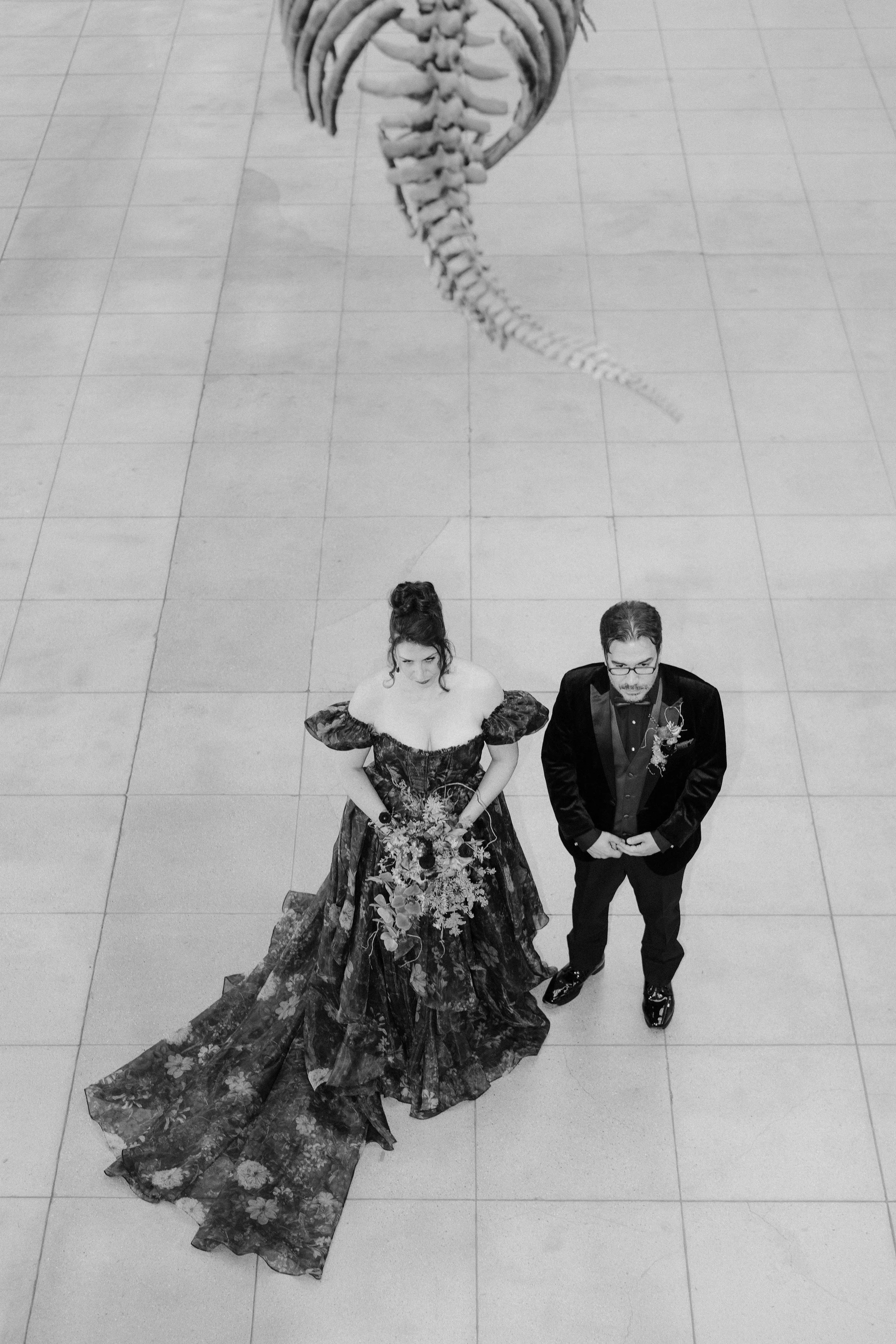 A bride and groom standing together outside in front of a white wall with a black gate, with an old brick building and greenery in the background. The bride is wearing a white strapless wedding gown with a veil, and the groom is wearing a dark suit with a yellow tie.