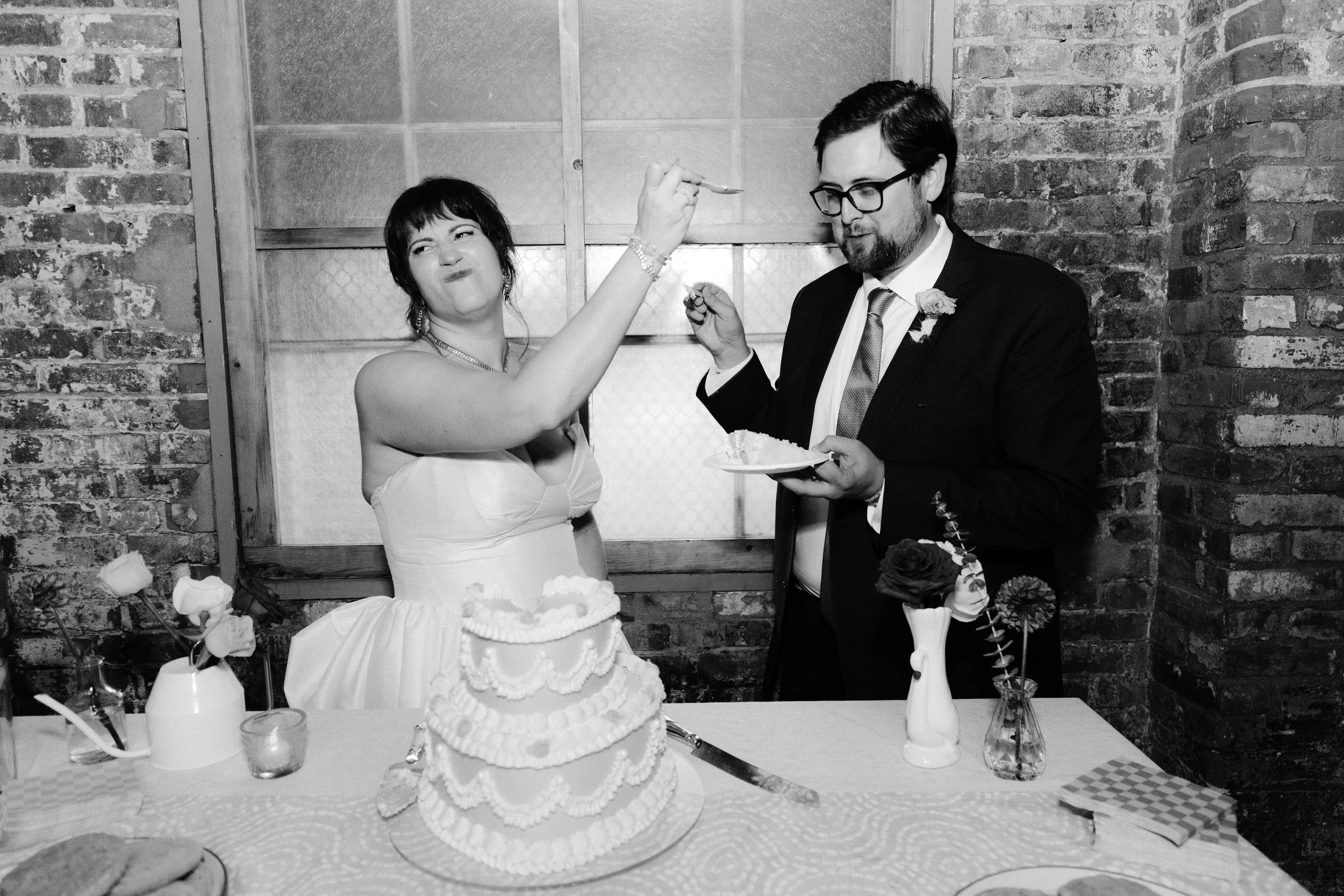 A woman in a strapless dress is feeding a man in a suit at a wedding reception. They stand behind a table with a large layered wedding cake, vases with flowers, and other items, against a brick wall background.