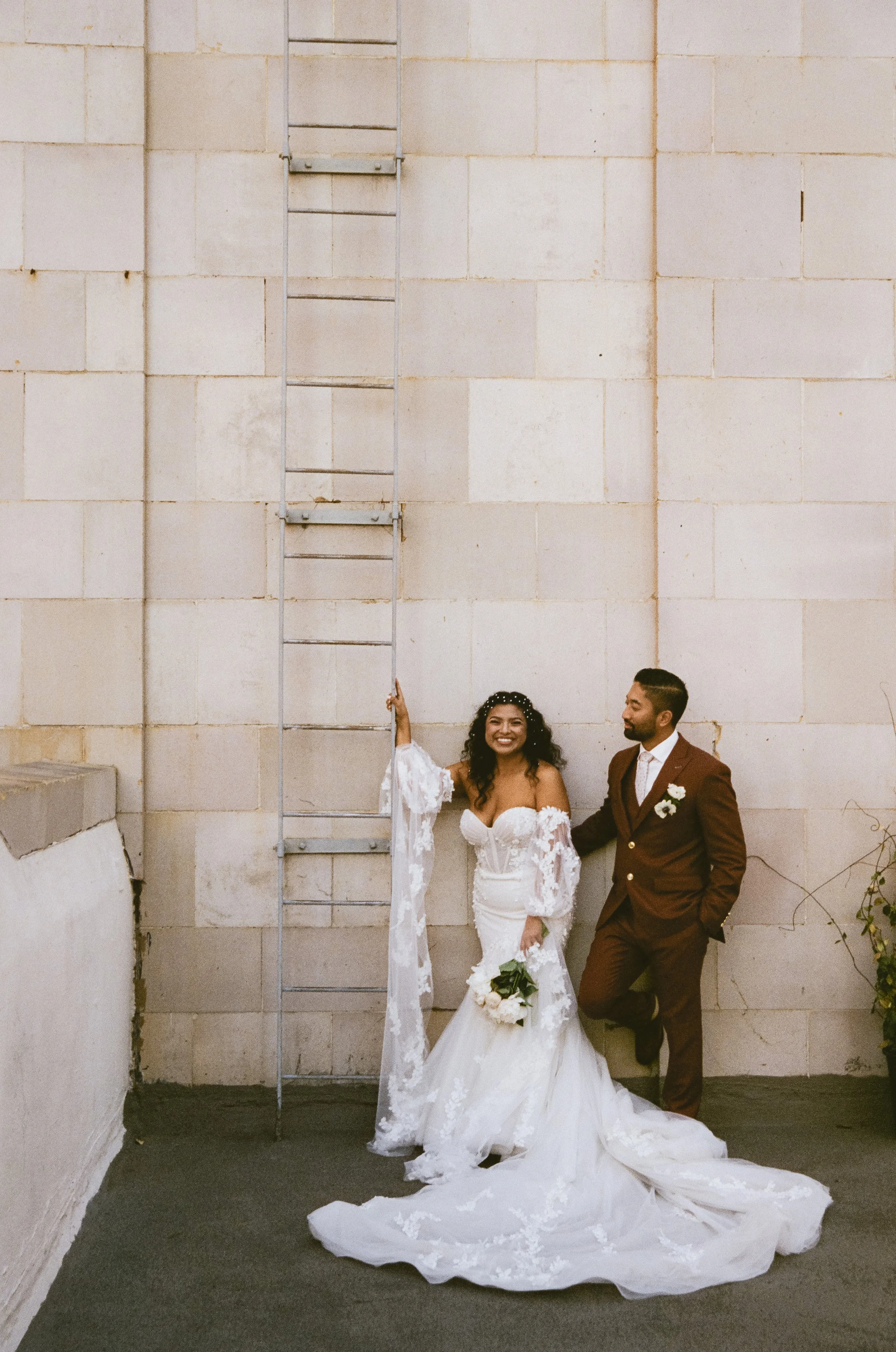 A bride and groom standing outdoors against a beige brick wall with a metal ladder. The bride is wearing a white wedding gown with lace details, holding a bouquet of flowers, and smiling. The groom is in a brown suit with a white shirt and boutonnier