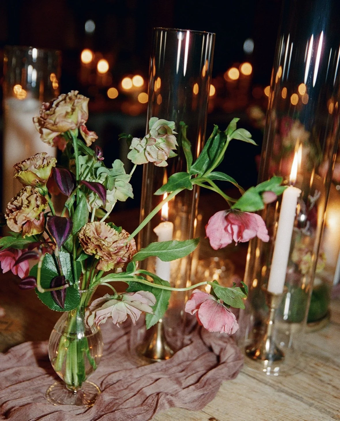 A floral arrangement with pink and purple flowers in a clear glass vase on a wooden table, with tall glass candle holders and lit candles in the background at a nighttime event.