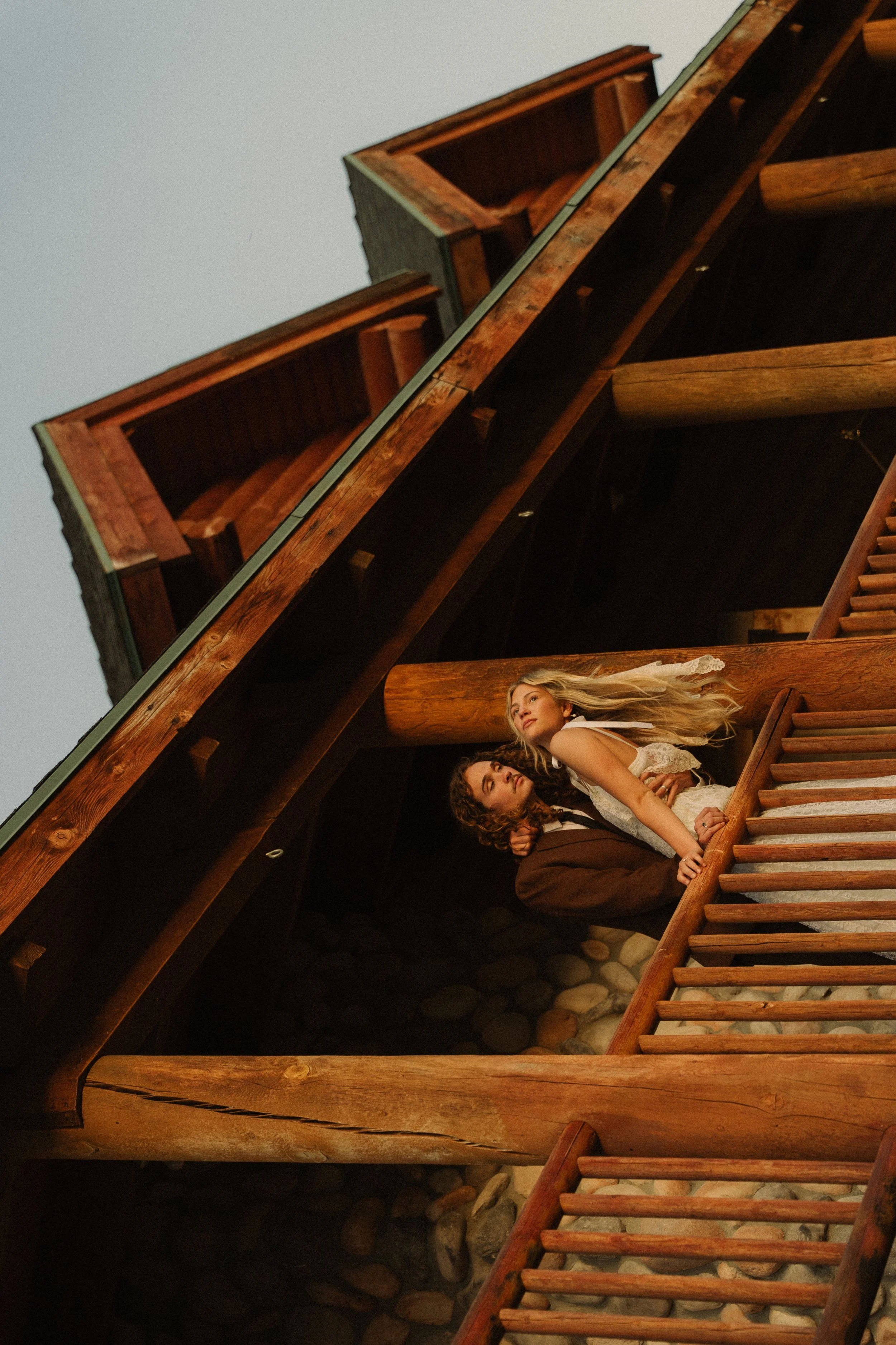 Two women are standing on a wooden staircase outside, near the edge of a building made of wood and stone, with one woman holding the other from behind. The woman in front has long blonde hair and is wearing a white lace dress, while the woman behind has curly hair and is wearing a dark suit.