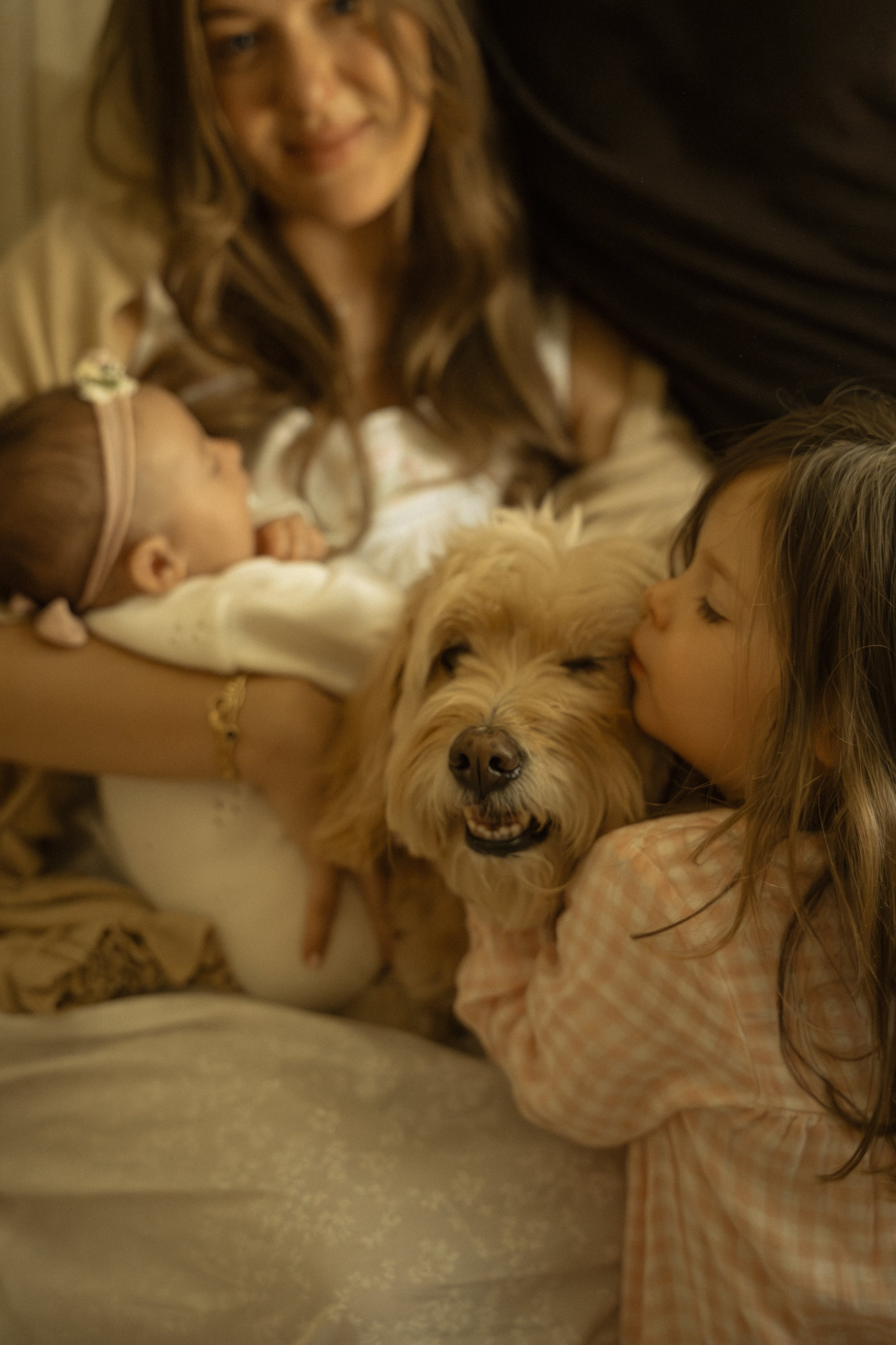 Two young girls are hugging and kissing a golden retriever puppy as a woman in the background smiles warmly.