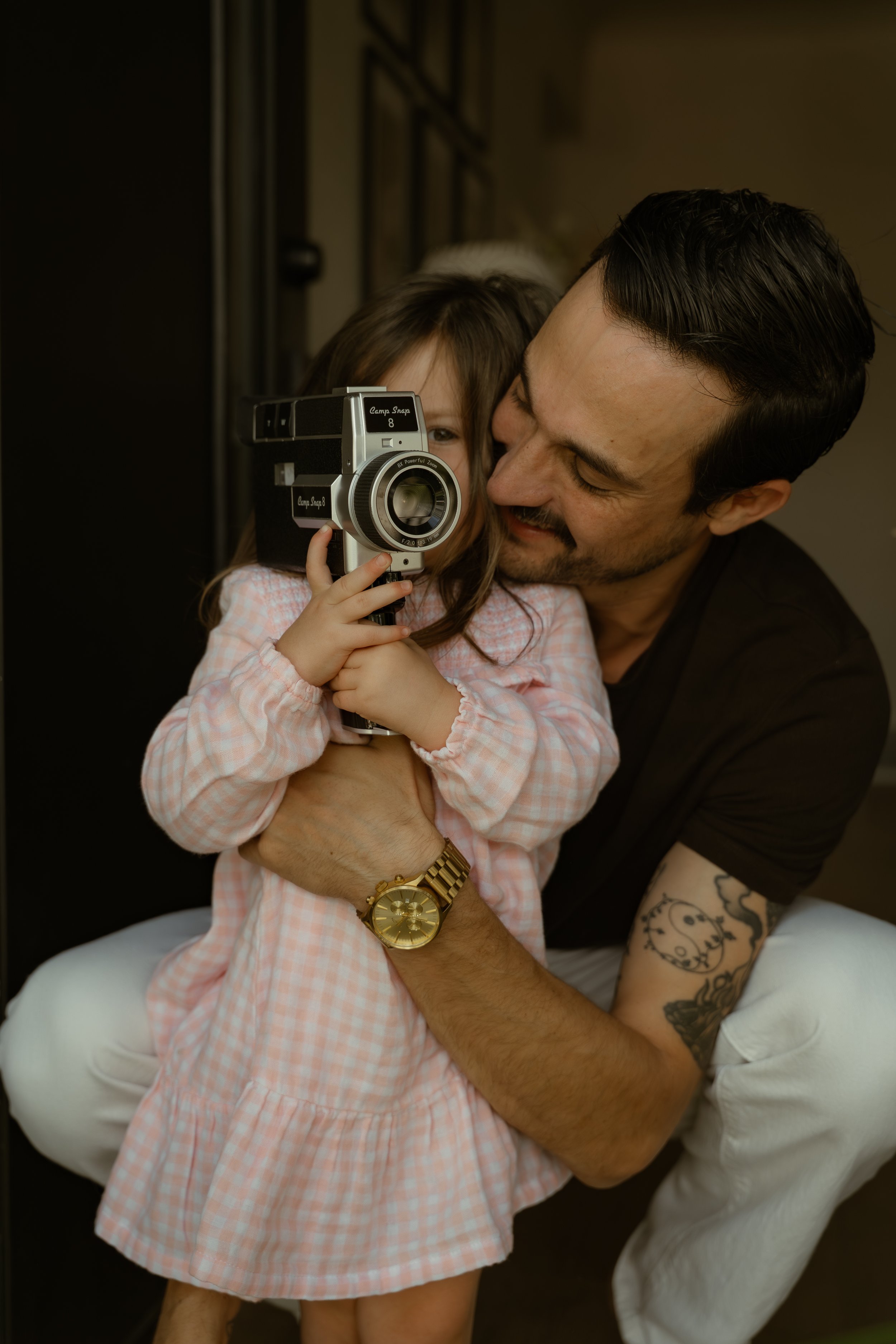 A man with tattoos on his arm and wearing a gold watch is hugging a young girl. They are both smiling and leaning close to each other. The girl is holding a vintage camera up to her face, covering most of it. The background is dark and out of focus.