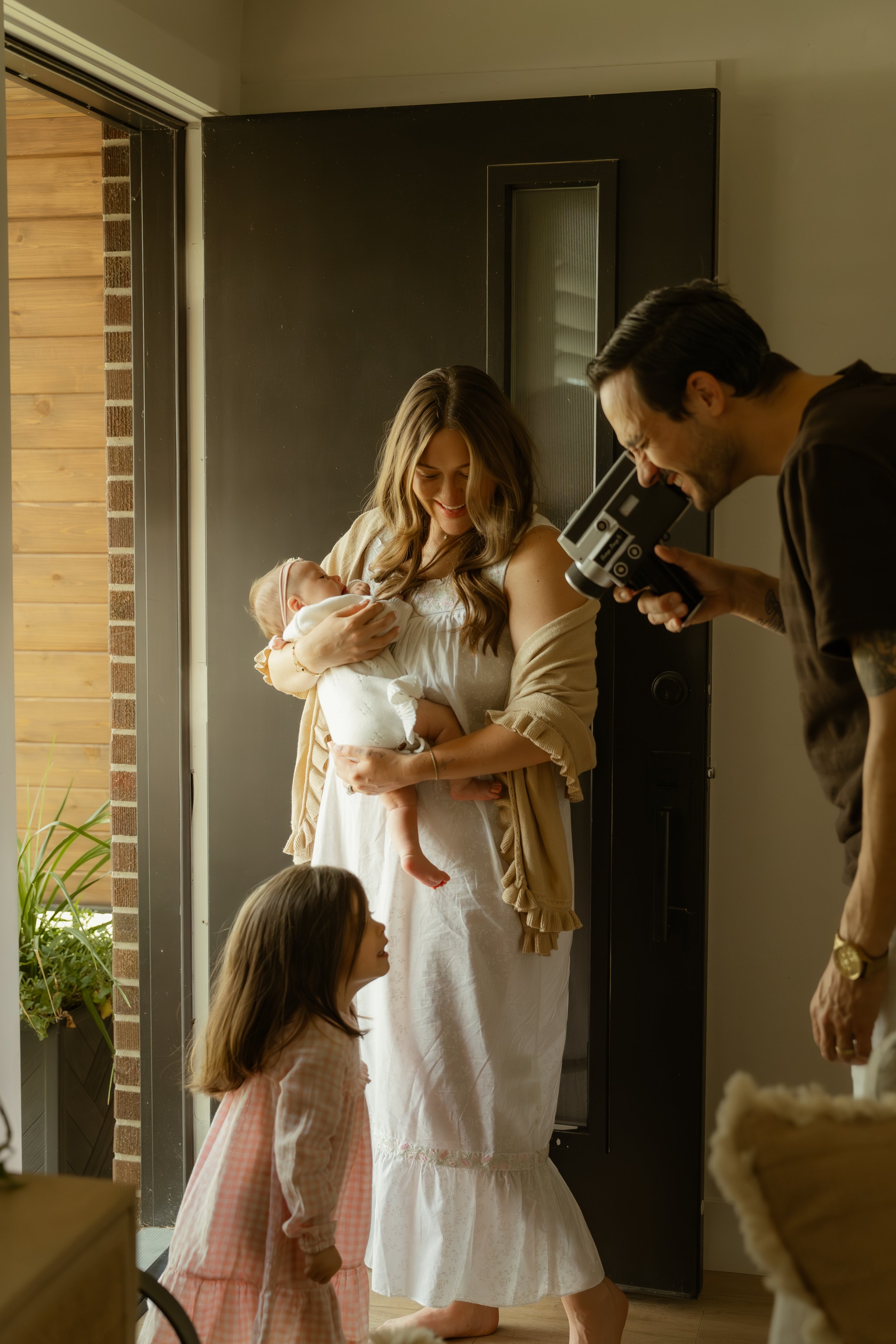 A woman holding a baby inside a house, with an older girl looking up to her, while a man films the scene with a camera.