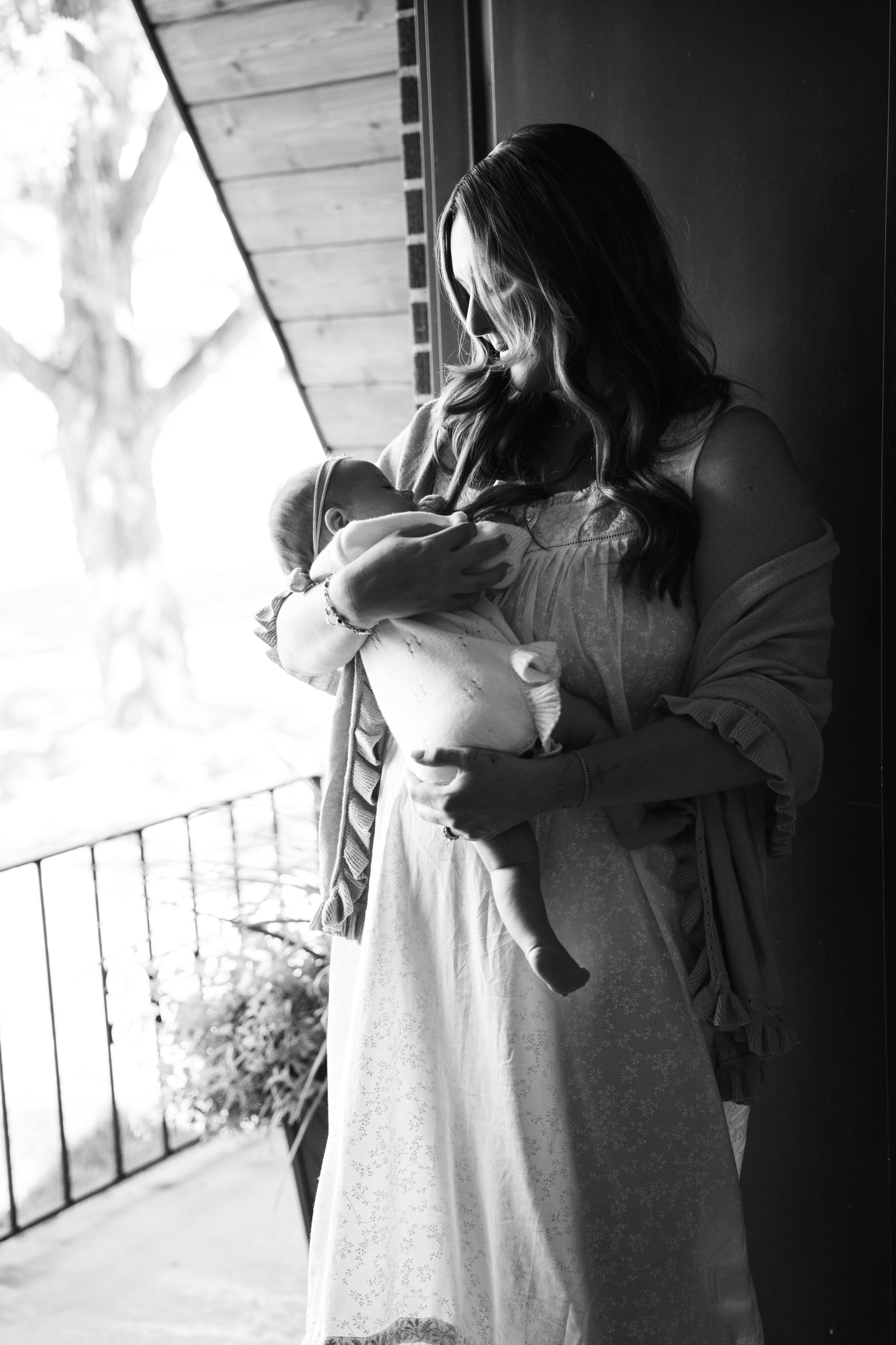 A woman with long hair, wearing a dress, holding a baby, standing on a balcony with a tree visible in the background.
