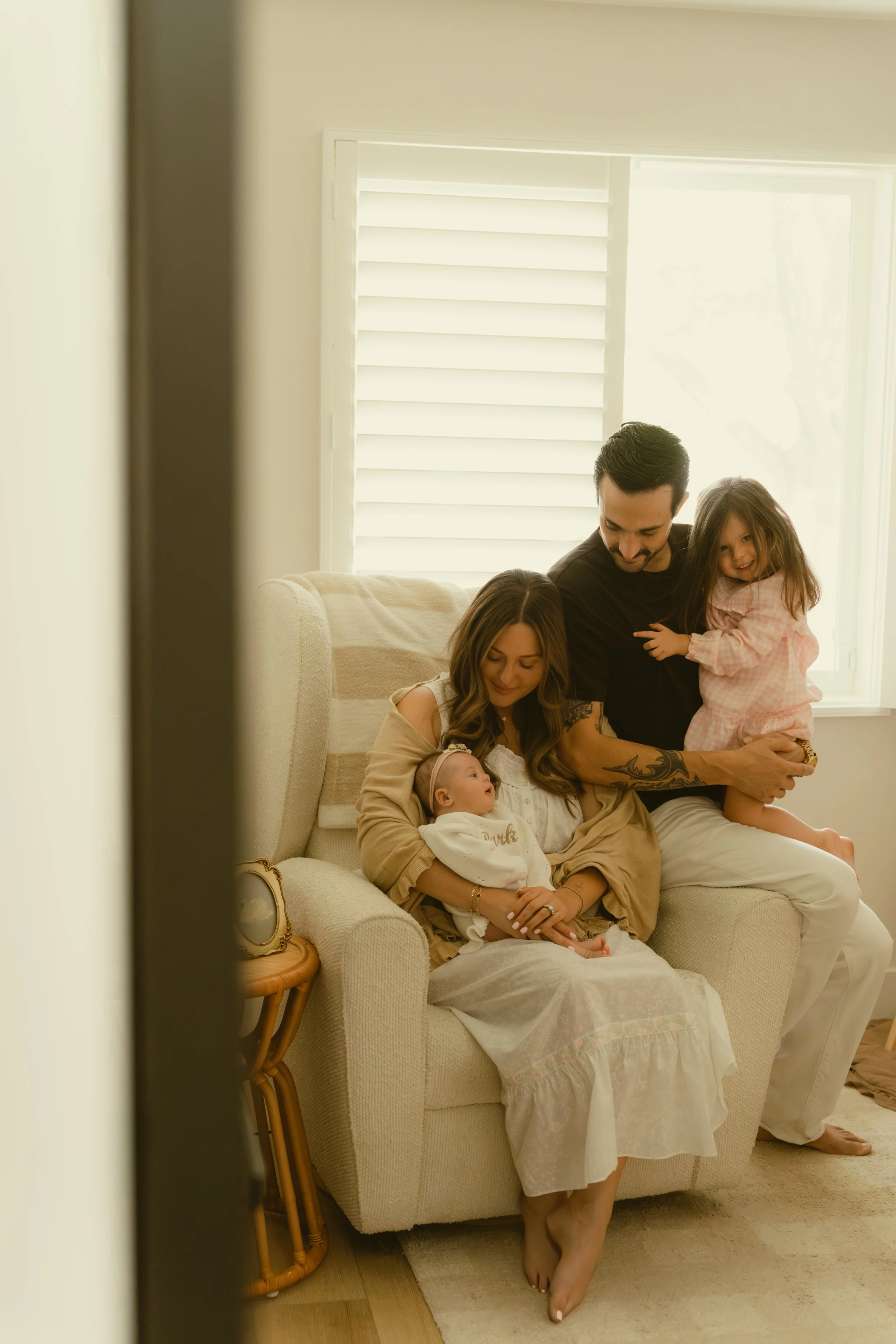 A family sitting on a beige couch in a bright living room, with a man, woman, and three young children, one girl standing on his lap and the youngest girl sitting on the woman's lap, all smiling and enjoying a moment together.