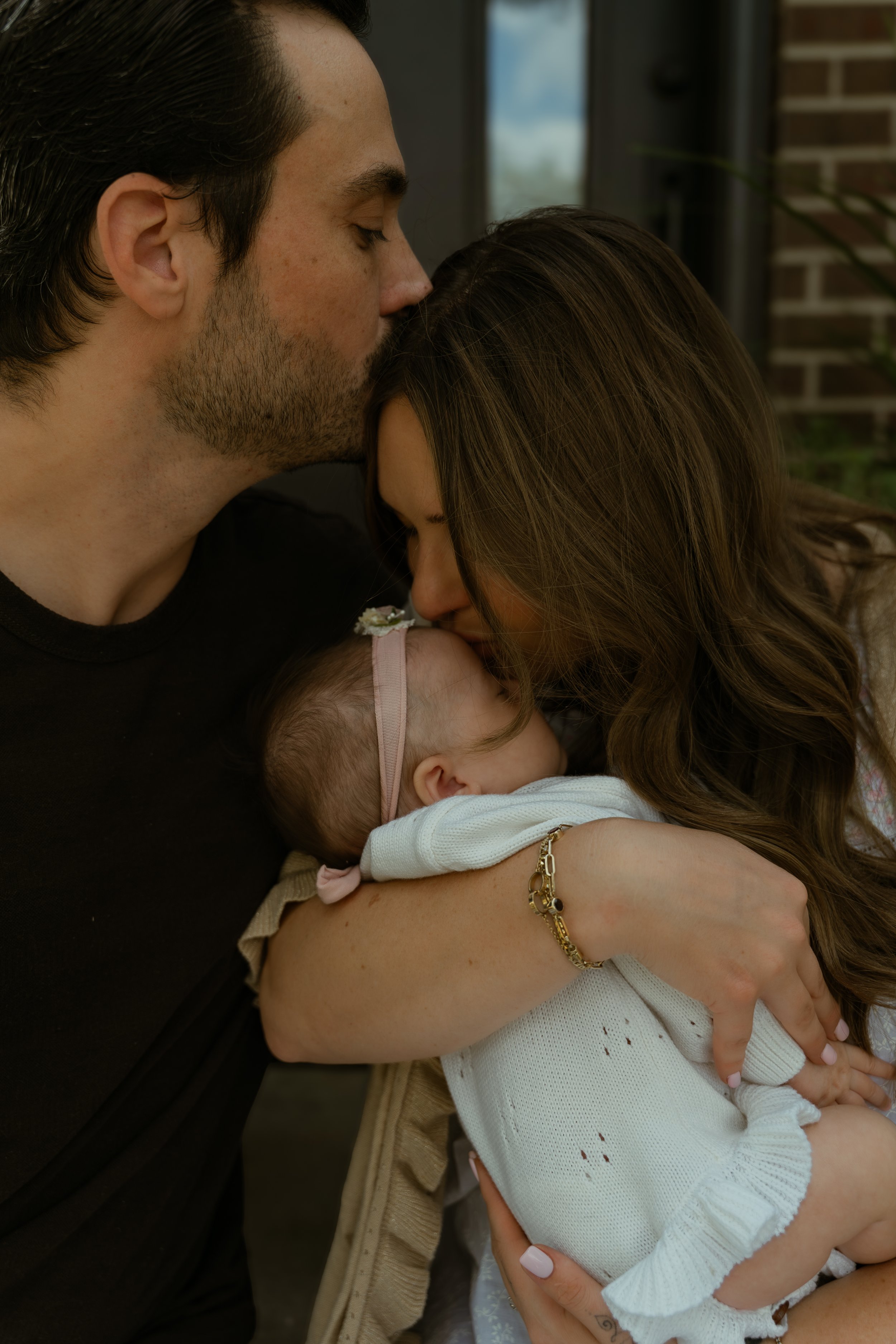 A couple holding their baby, the man is kissing the woman on her forehead while she is holding the sleeping baby, near a window with brick wall view.