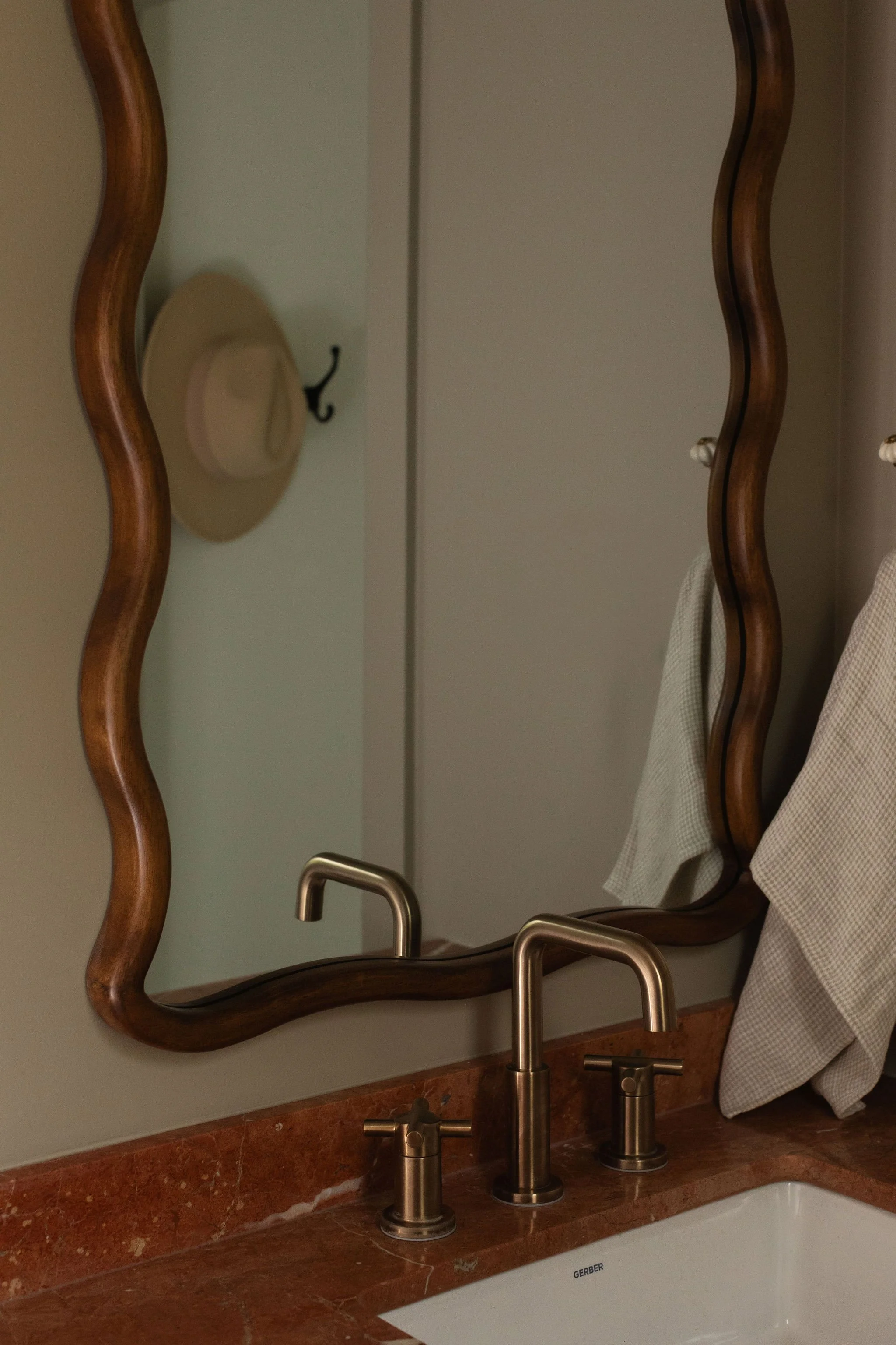 Detail of a wavy wood-framed bathroom mirror, aged brass faucet and red marble countertop.