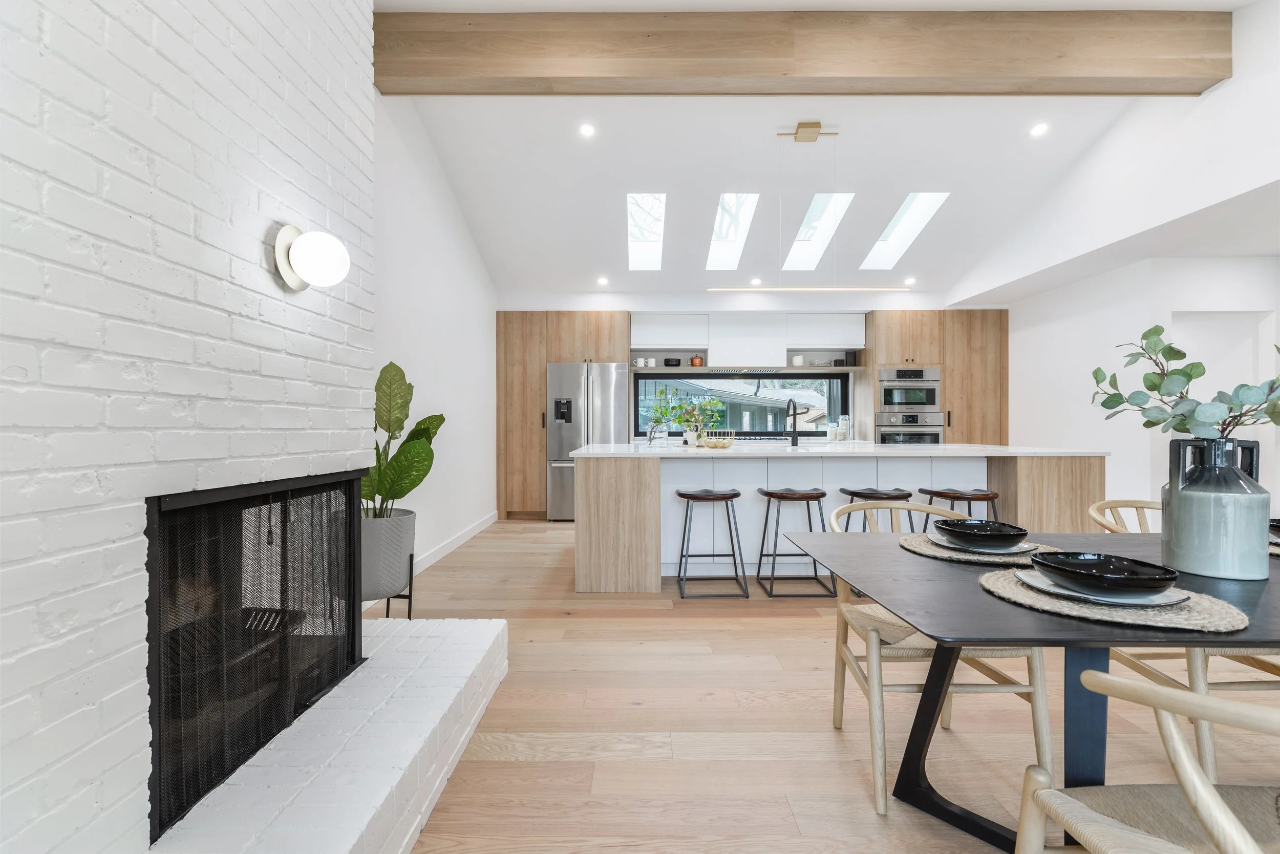 Open kitchen with vaulted ceiling and skylights. white oak floors and cabinetry. White brick fireplace