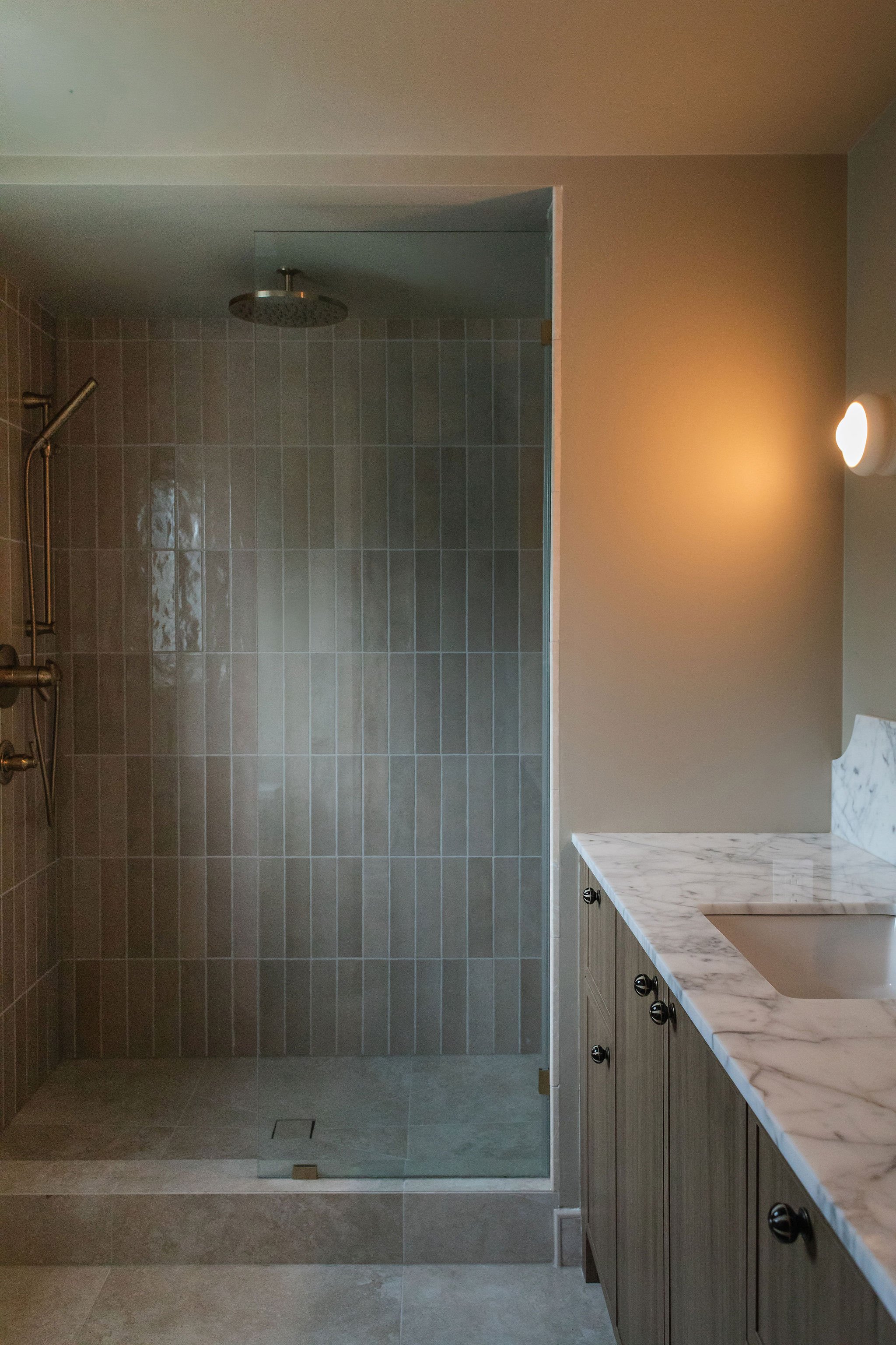 Bathroom renovation with a glass shower enclosure, vertical taupe tile, marble vanity countertop and warm brass fixtures.