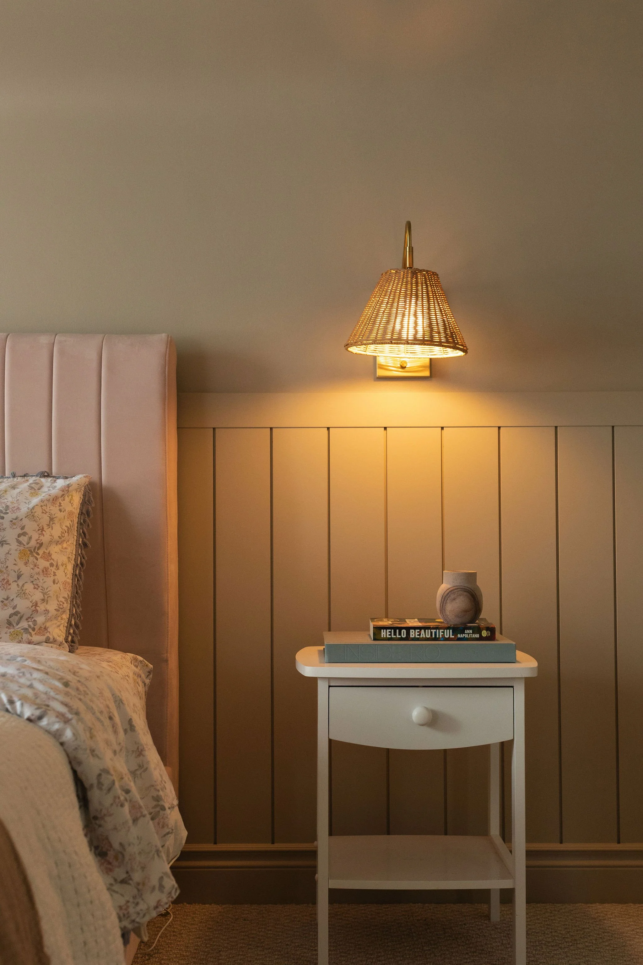 Cottage bedroom featuring a blush upholstered headboard, floral bedding, sage-green wall panelling and warm bedside lighting.