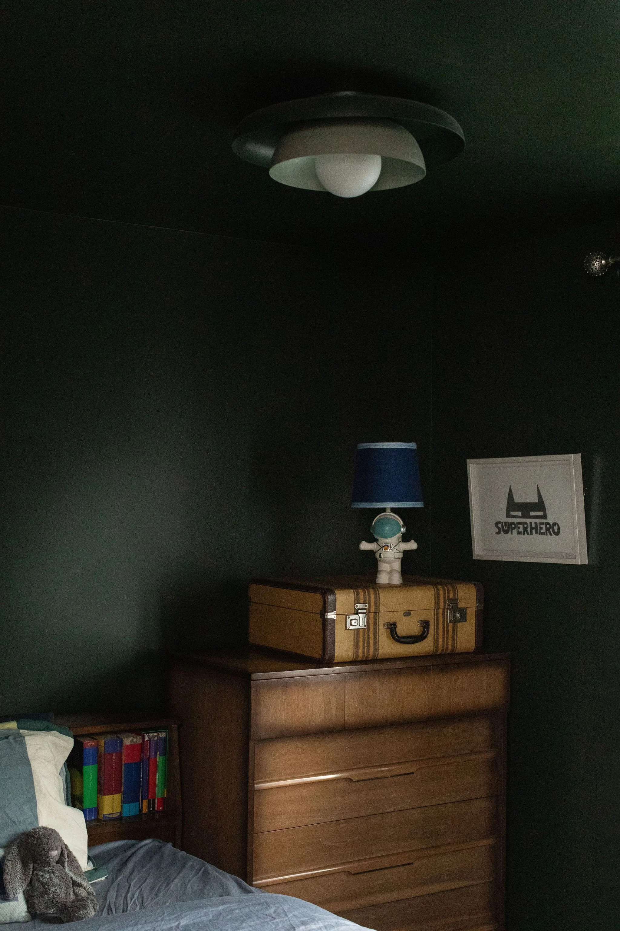 Moody bedroom featuring deep green walls, a vintage wood dresser, a classic suitcase and playful collected decor.