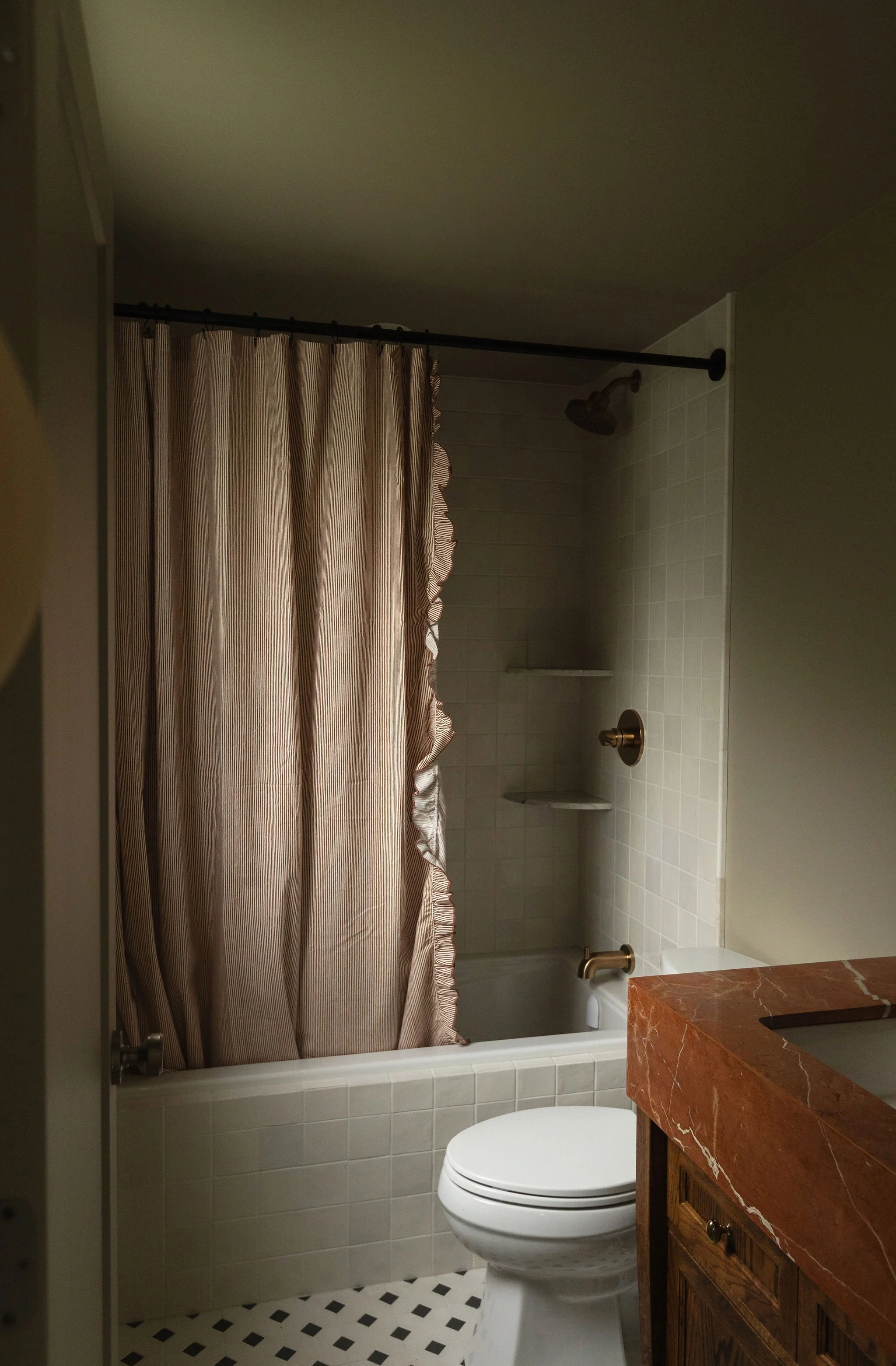 Cottage bathroom featuring a white bathtub, textured shower curtain, black and white floor tile and a red marble vanity countertop.