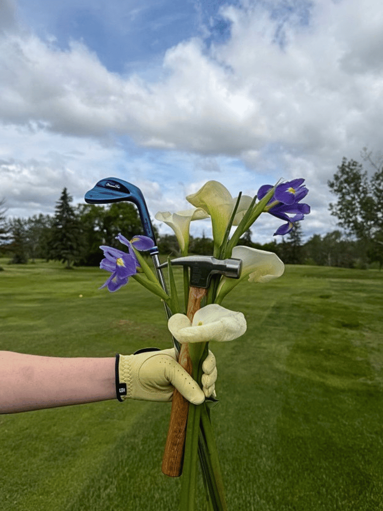 Hand wearing a golf glove holding a bundle of golf clubs and flowers on a golf course with trees and cloudy sky in the background.